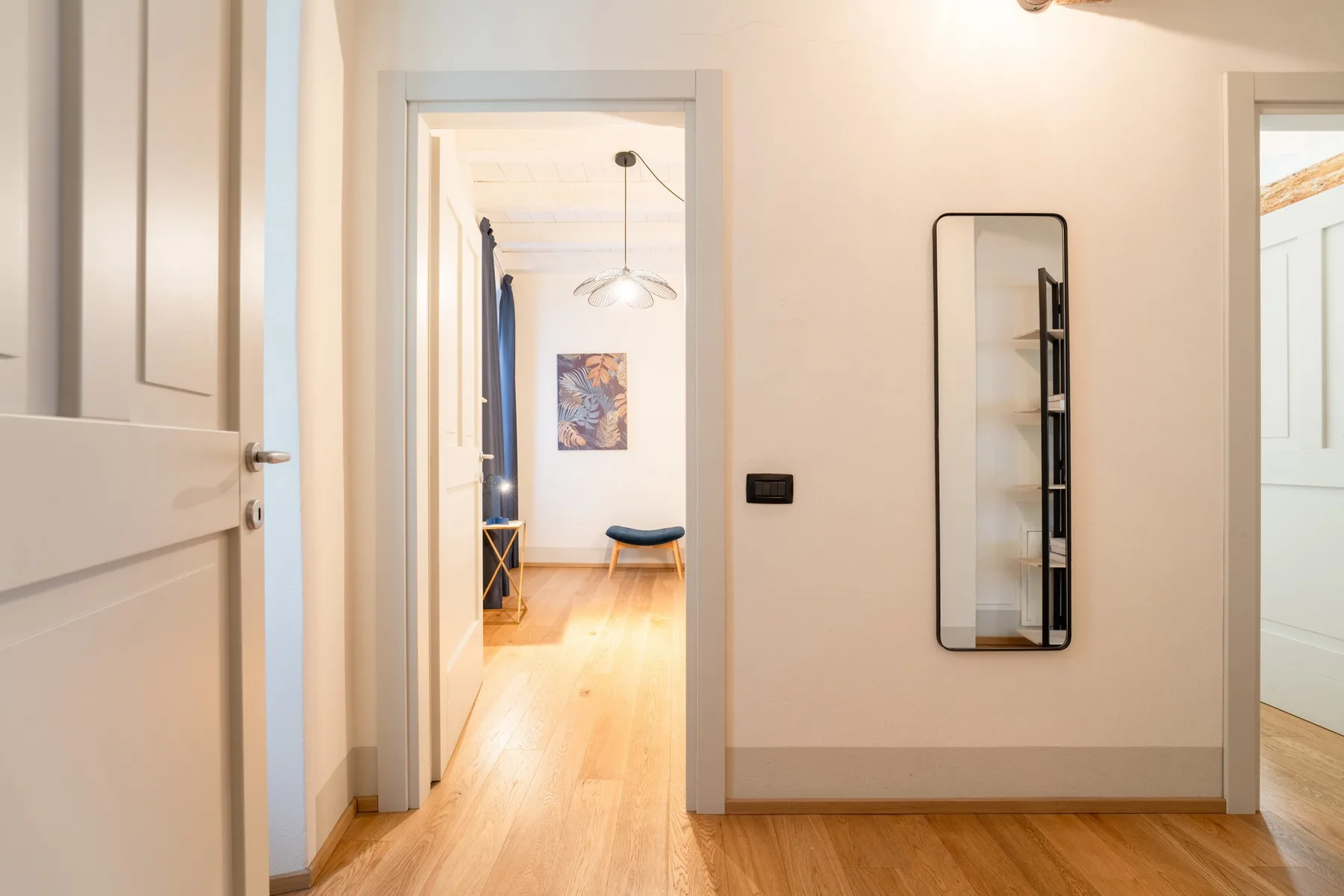 Un pasillo luminoso con suelo de madera, puertas abiertas y un espejo en la pared.
