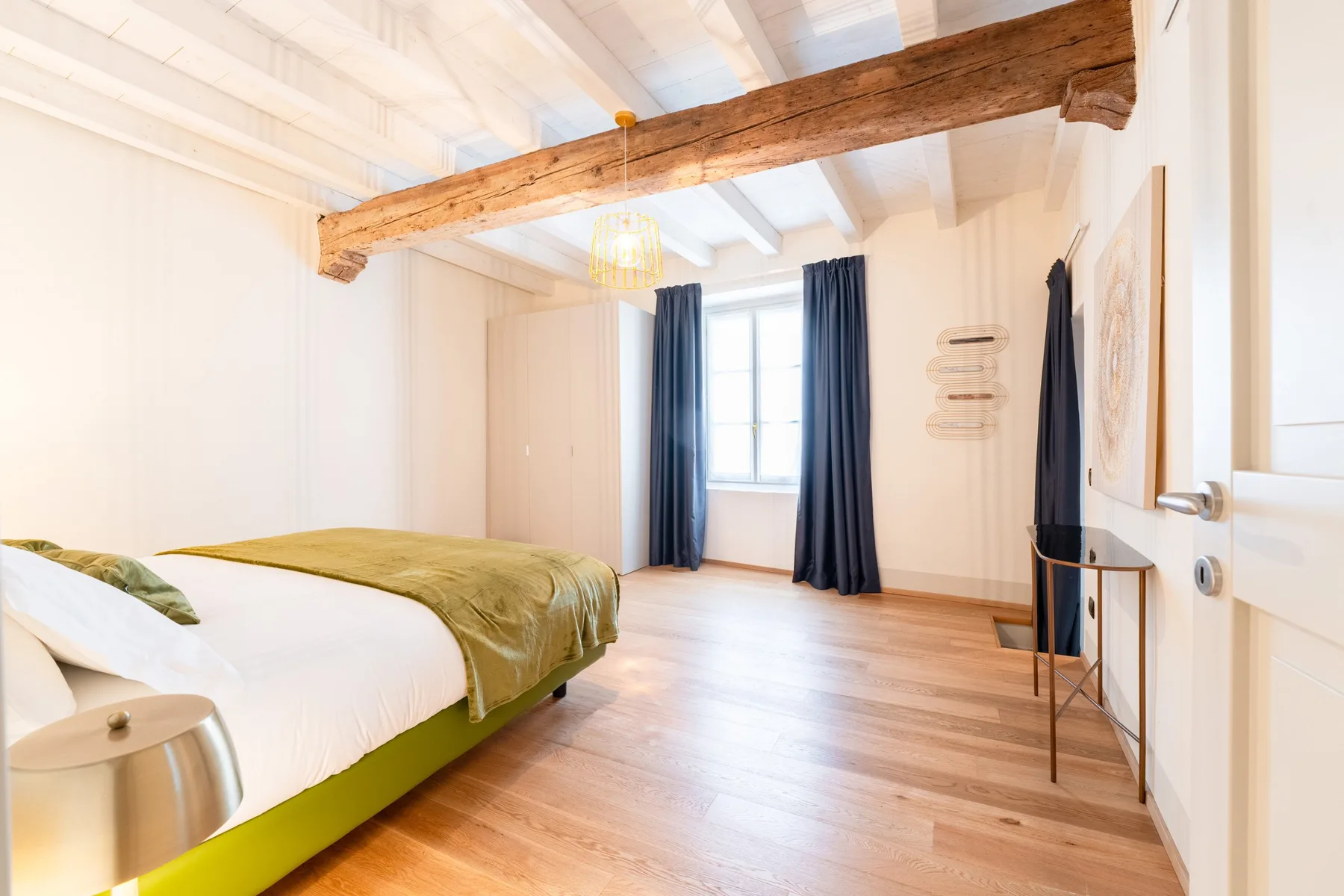 Un dormitorio luminoso con una cama, cortinas oscuras y vigas expuestas en el techo.