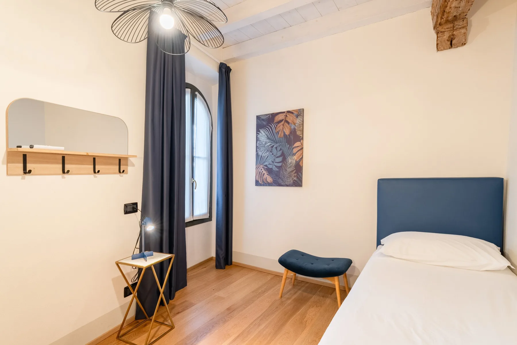 Un dormitorio con una cama, un pequeño asiento tapizado y una ventana alta con cortinas.