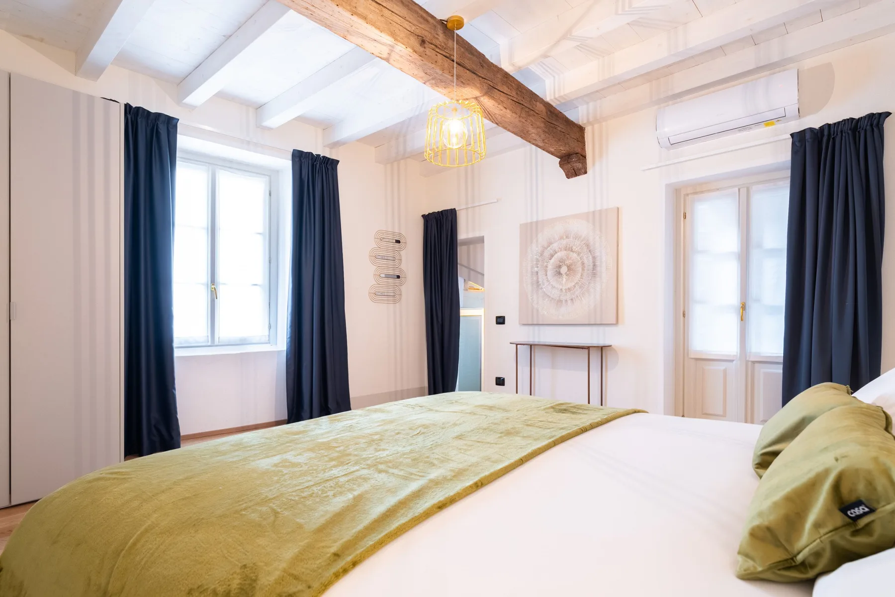 Un dormitorio luminoso con una cama, cortinas oscuras y vigas expuestas en el techo.