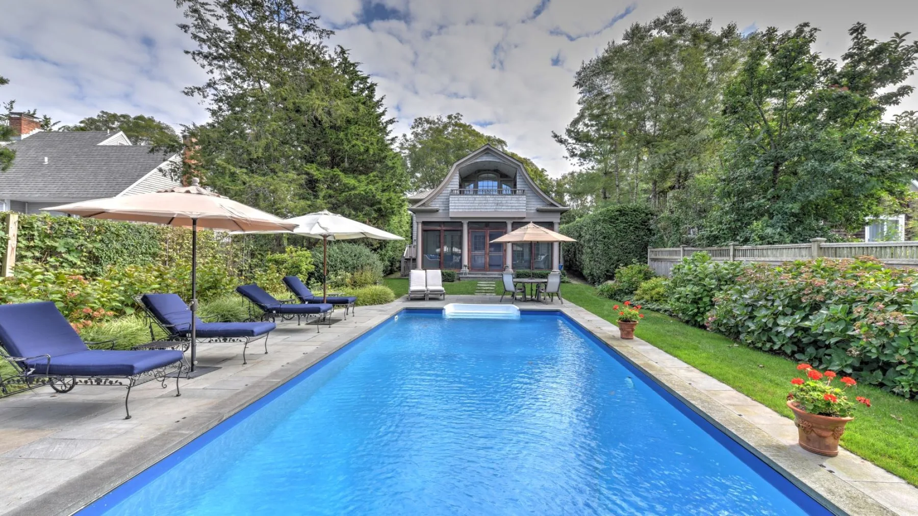 Una piscina en el patio trasero conduce hacia una casa, con asientos y sombrillas junto al patio.