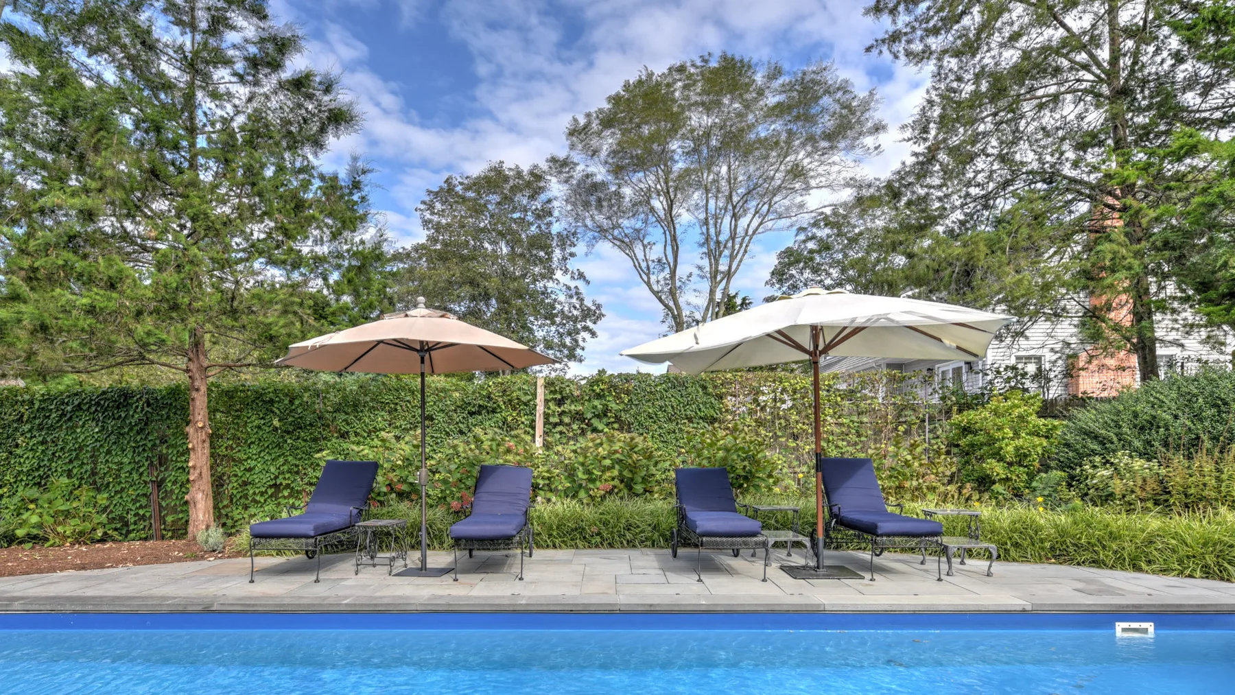Un patio junto a una piscina tiene asientos reclinables bajo dos sombrillas grandes.
