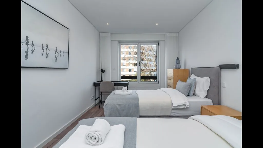 Un dormitorio luminoso con dos camas y una ventana con vistas a edificios cercanos.