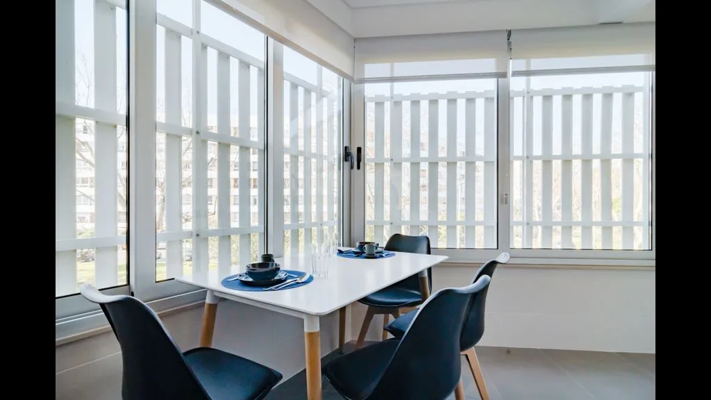 Un comedor con una mesa blanca y asientos oscuros junto a grandes ventanales.