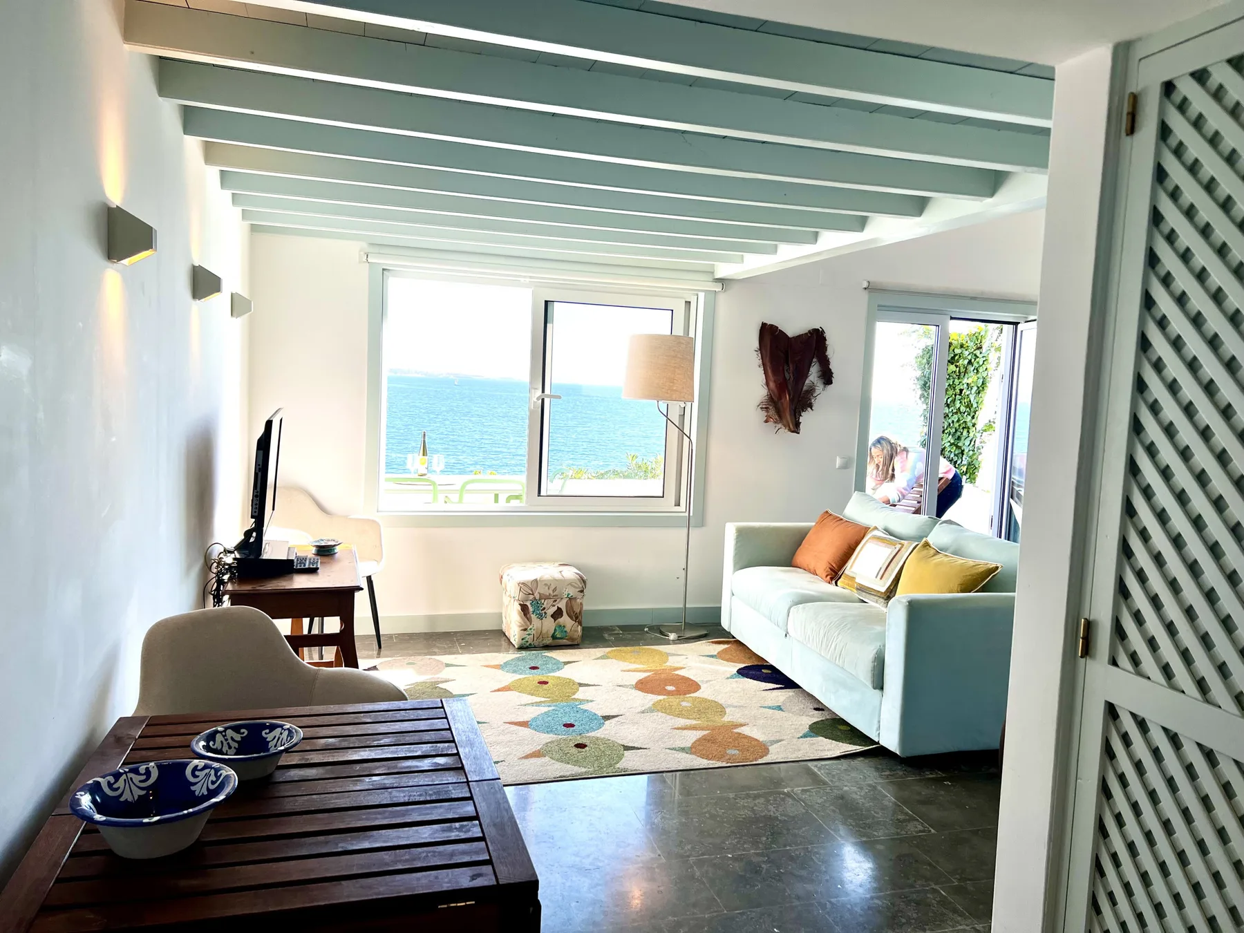 A bright living room with a pale sofa, colorful rug, and windows overlooking the sea.