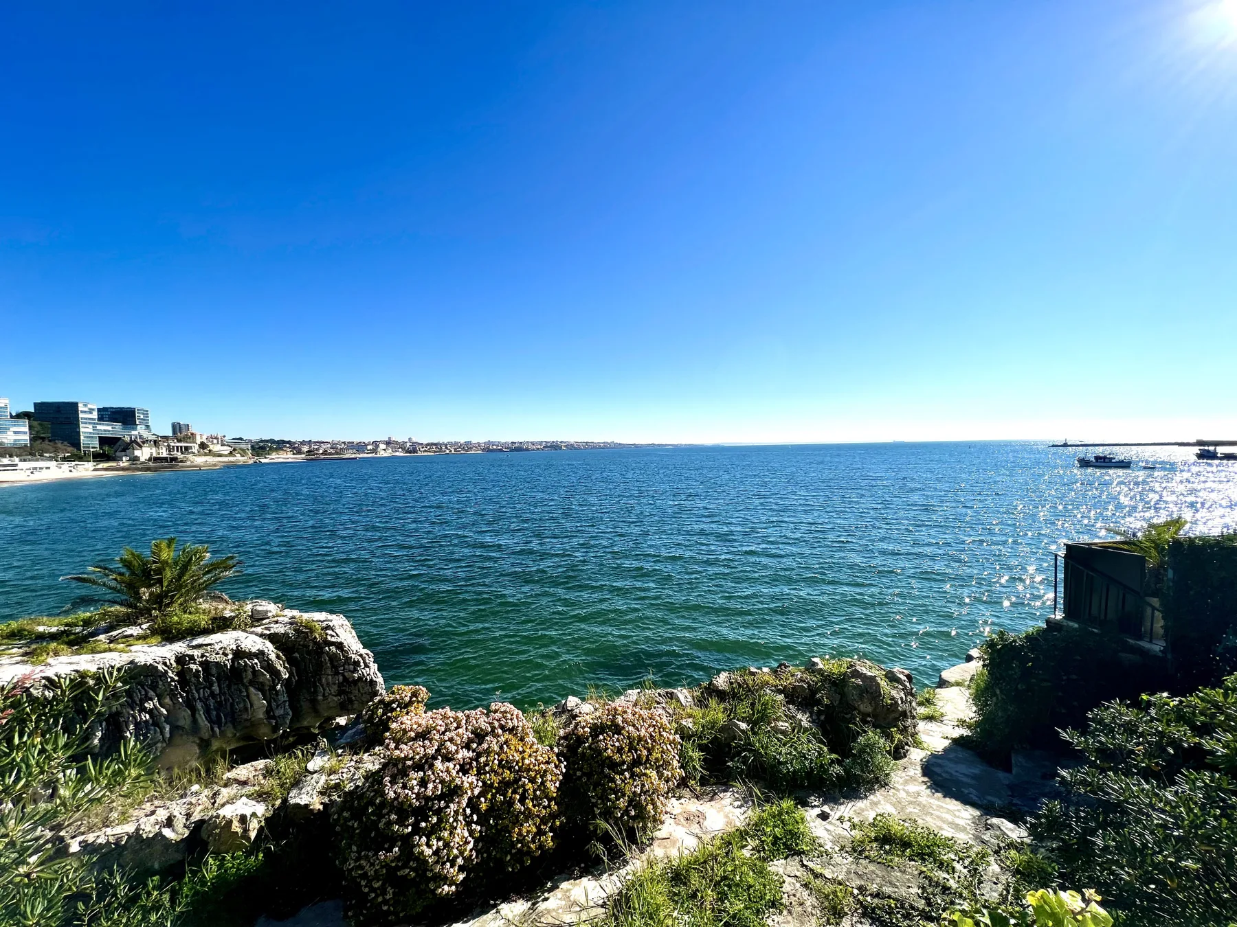 A seaside view shows blue water, a rocky shoreline, and distant buildings.