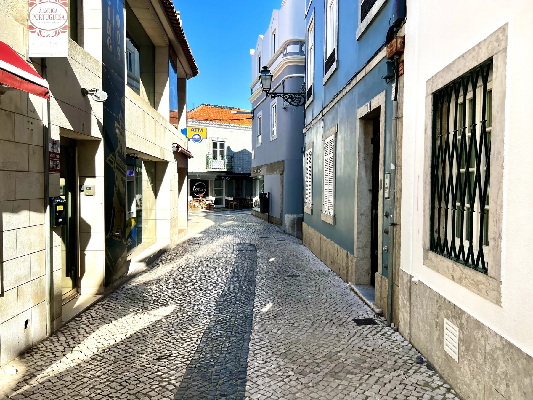 A narrow cobblestone street runs between closely spaced buildings in bright sunlight.