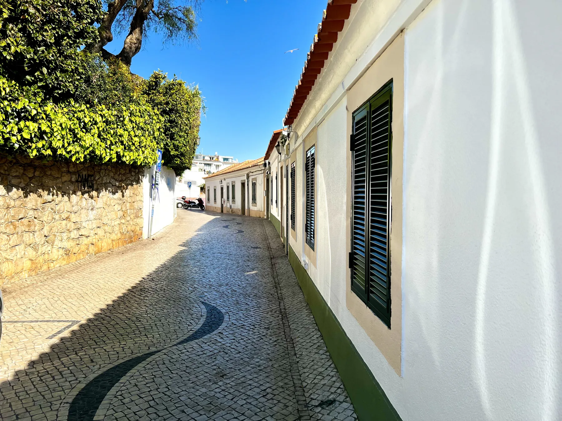 A cobblestone street runs past white buildings and a stone wall beneath a clear blue sky.