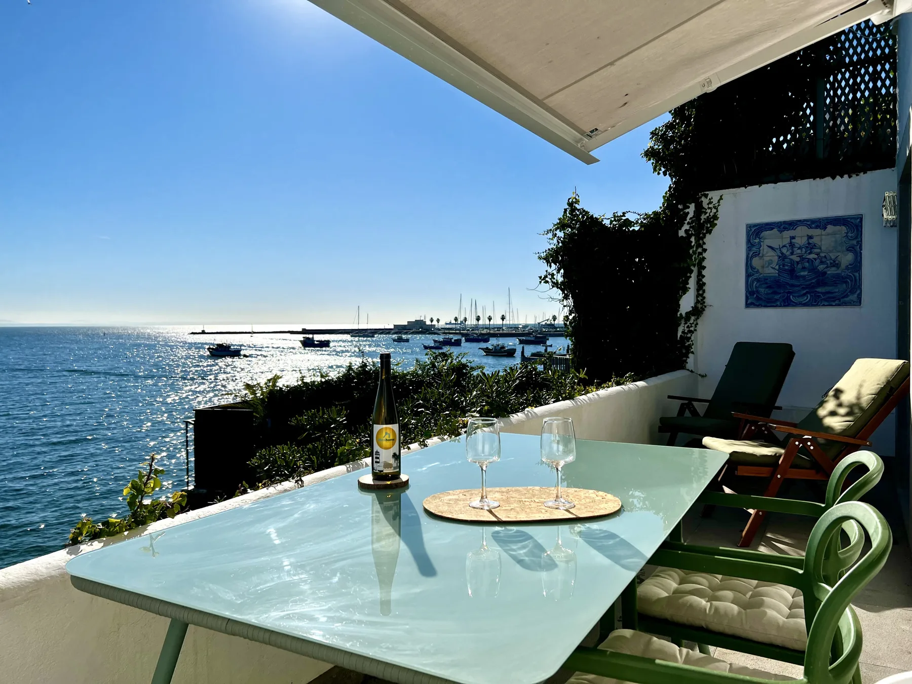 A seaside terrace with a glass-topped dining table and green seating overlooks the water.