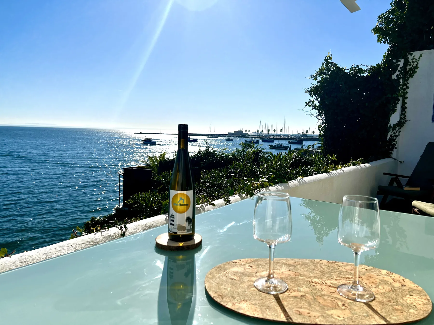 A seaside terrace table overlooks the water, with a bottle and two glasses.