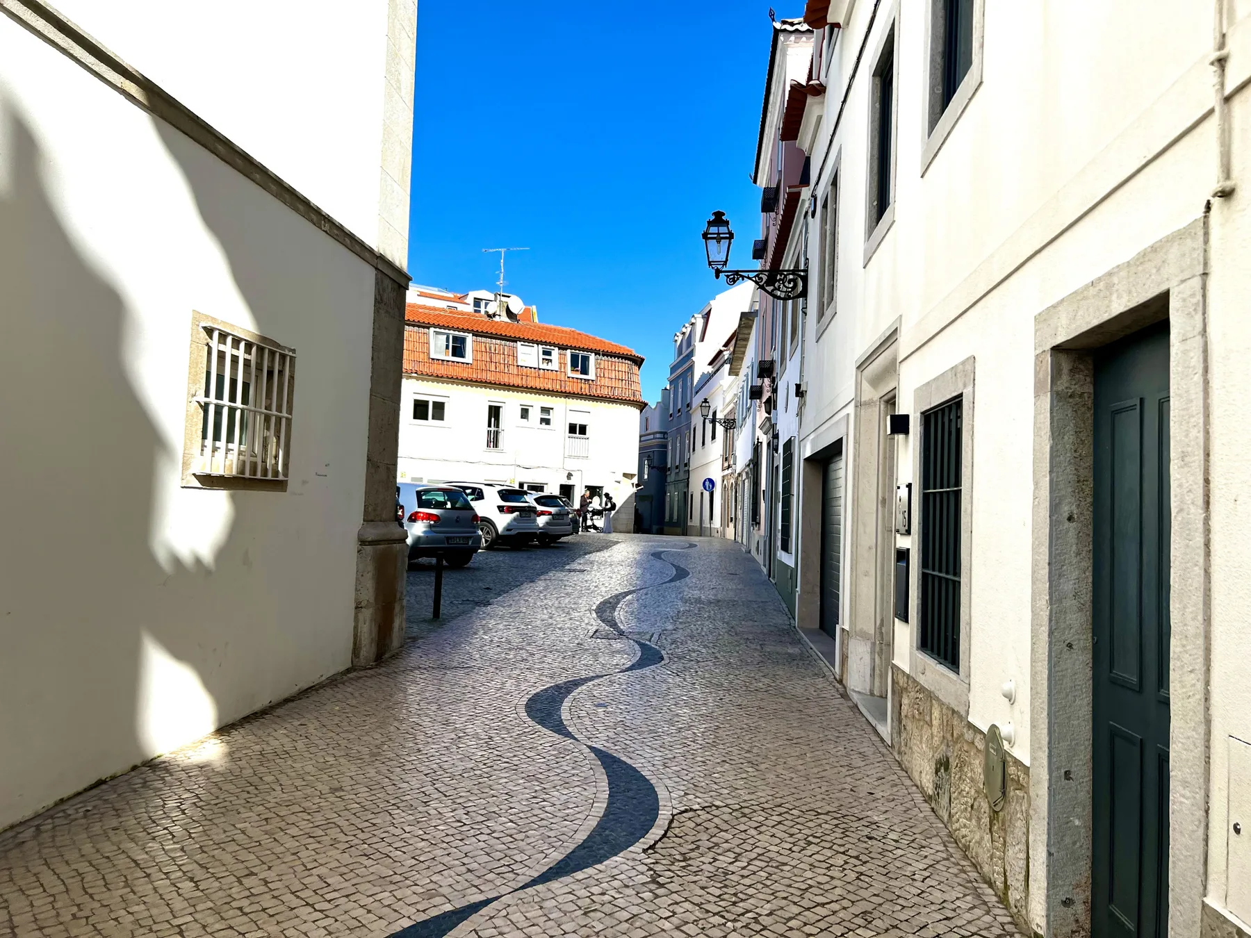 A narrow cobblestone street runs between white buildings under a bright blue sky.