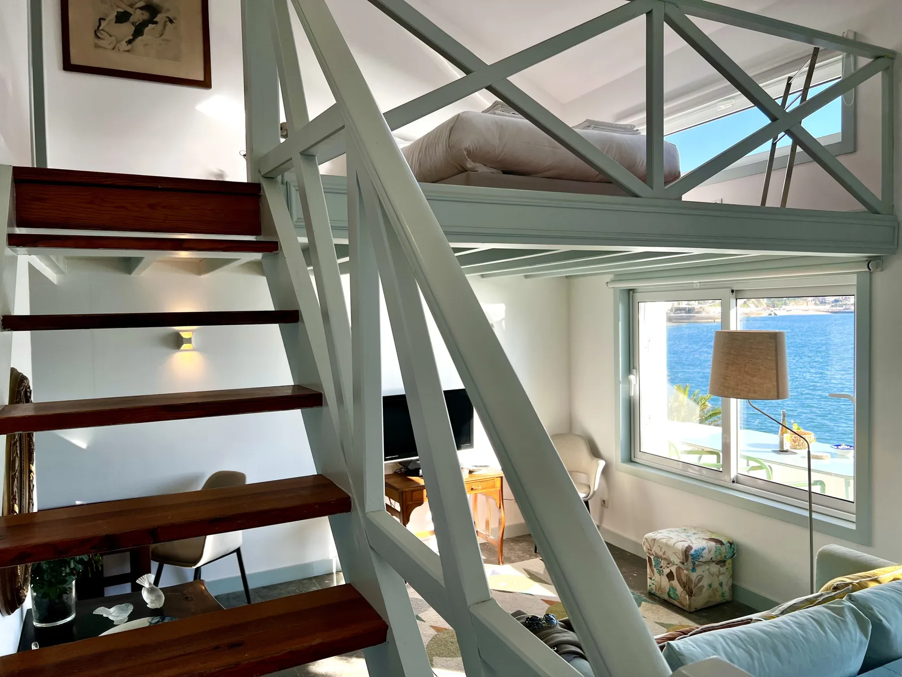A bright loft living area with a staircase and large windows overlooking the sea.