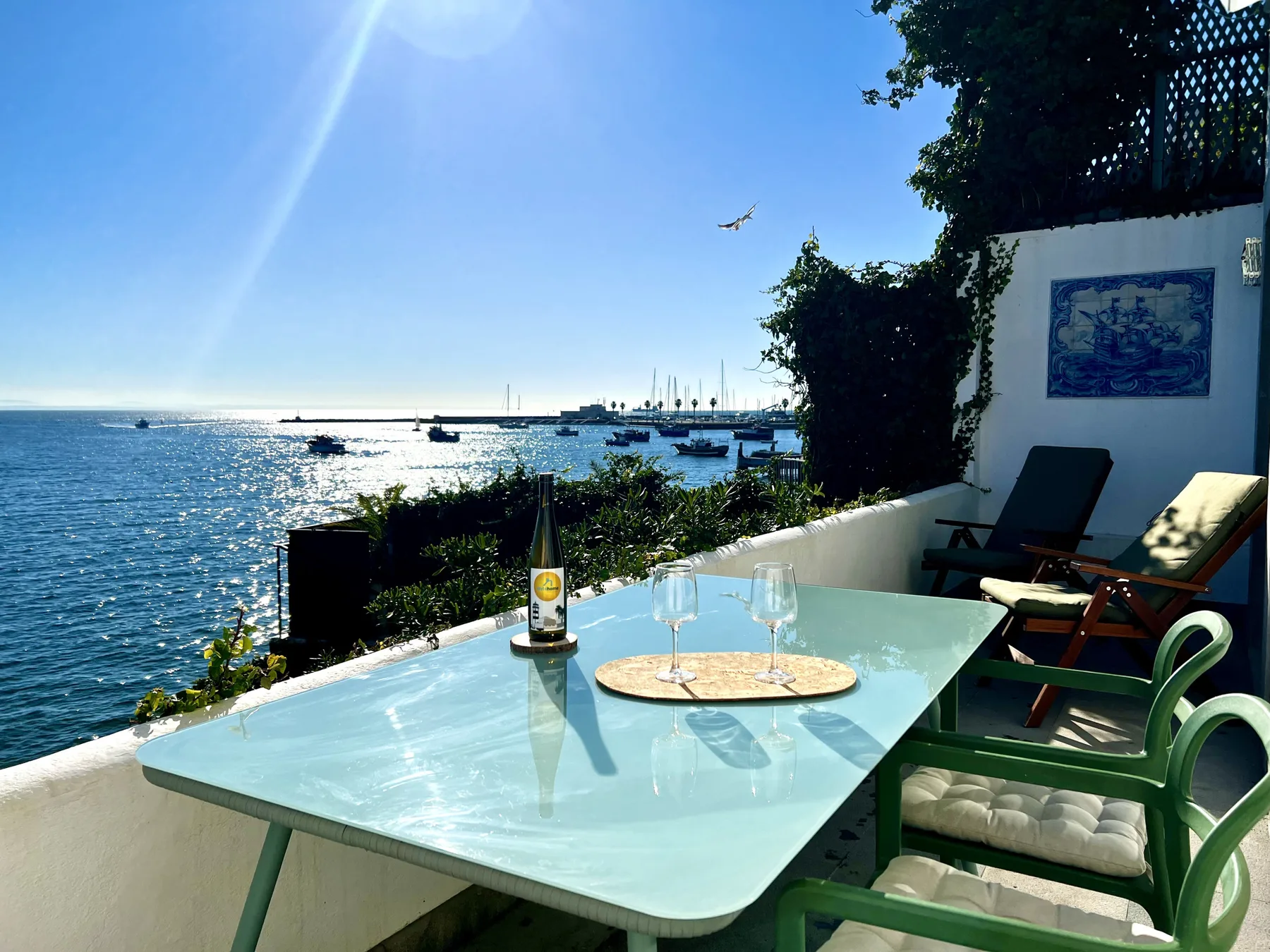 A seaside terrace has a glass table and green seating overlooking the water.