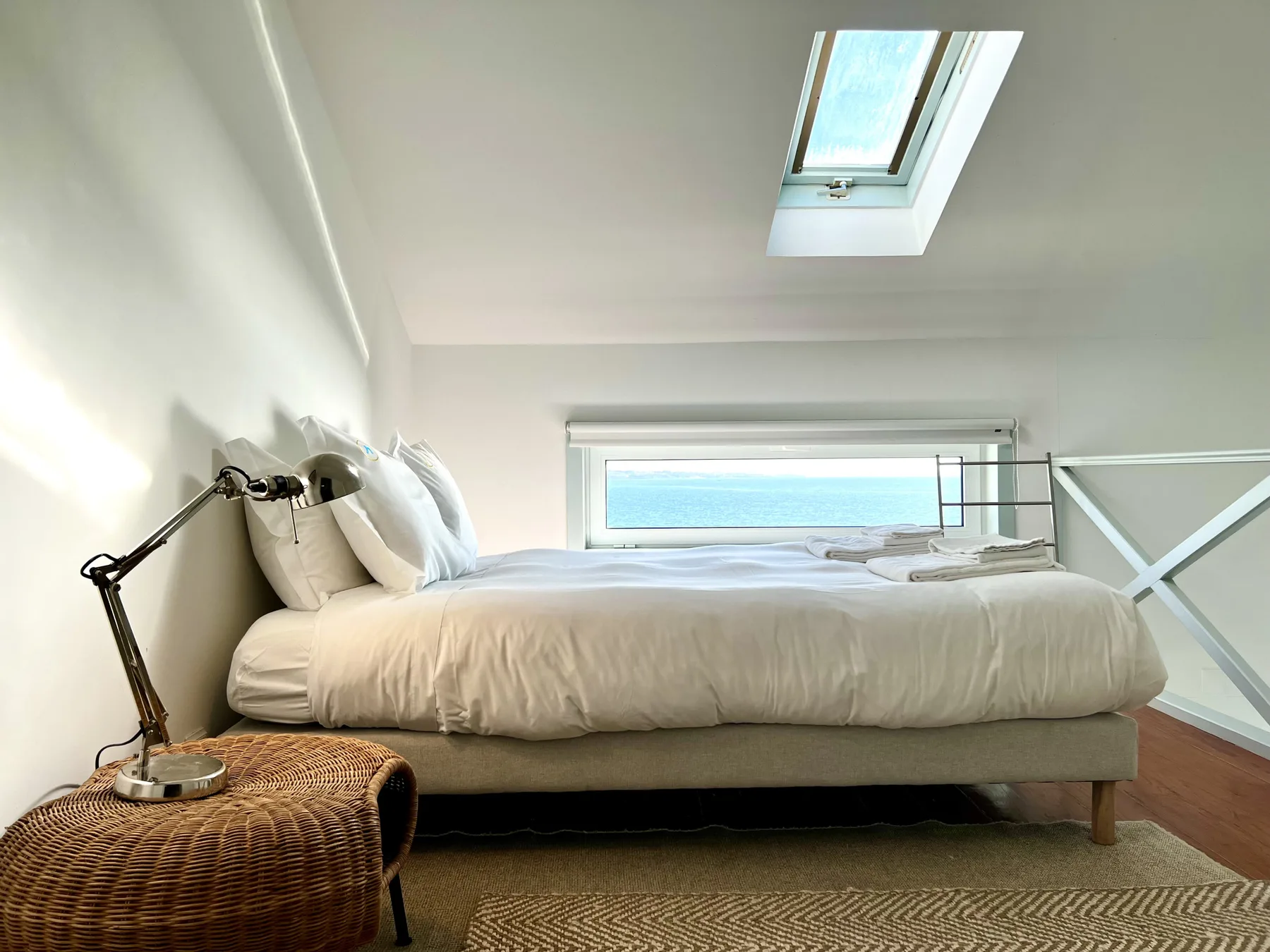 A bright bedroom with a bed beneath a skylight and a window overlooking the sea.