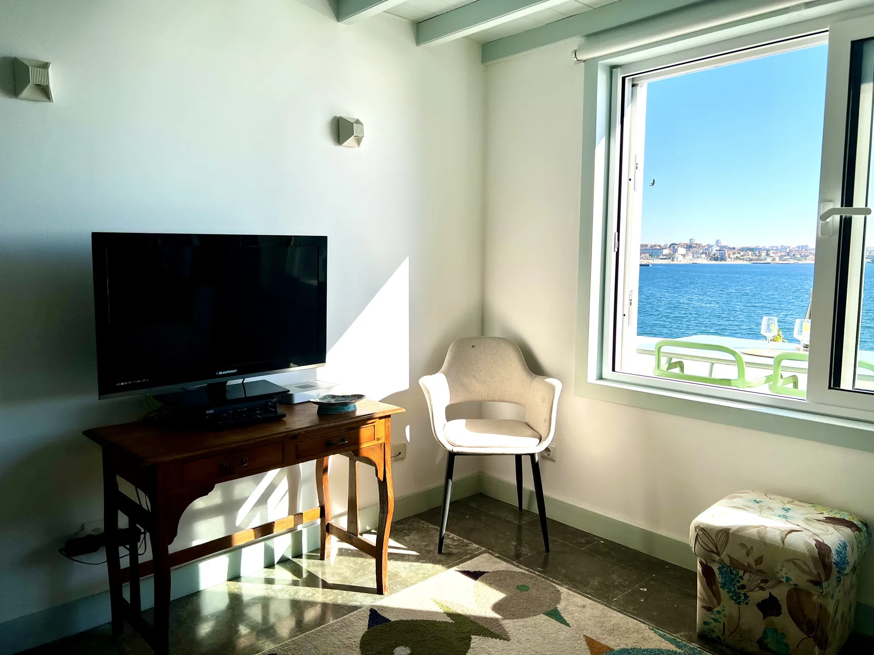 A sunlit living area has seating beside a large window overlooking the sea.