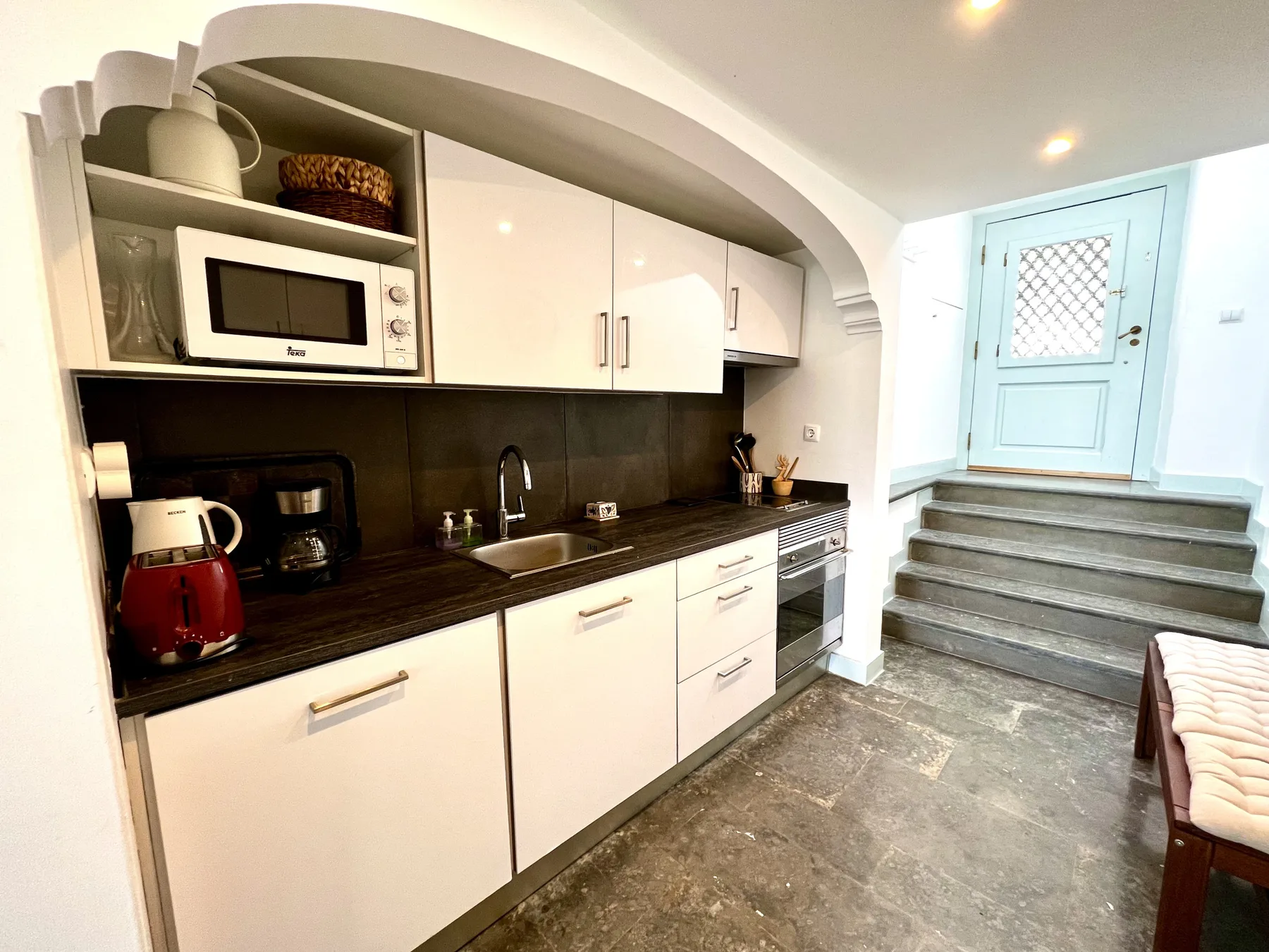 A compact kitchen with white cabinets and a dark countertop beside a short staircase.