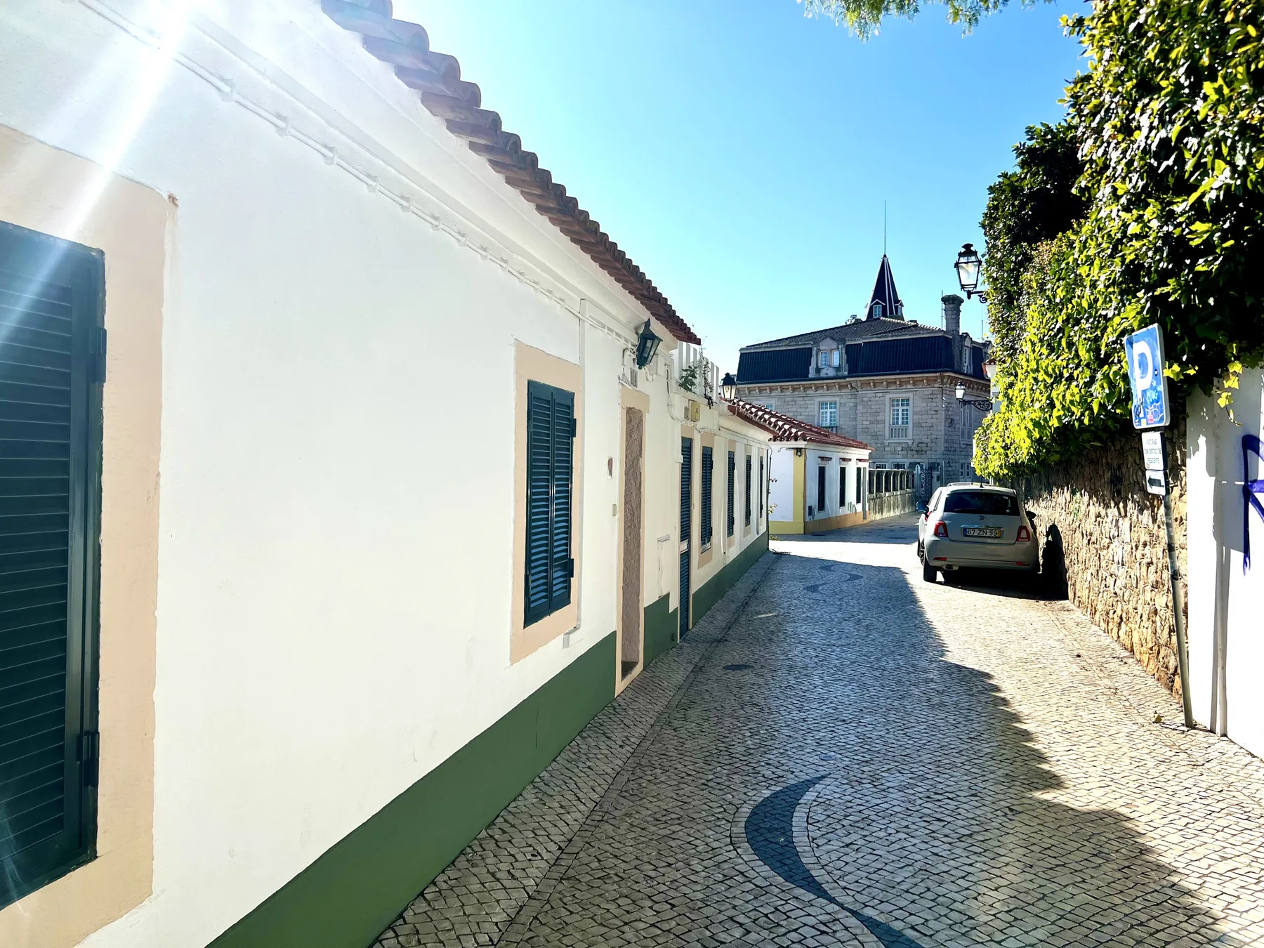 A narrow cobblestone street runs beside a row of white buildings with green trim.