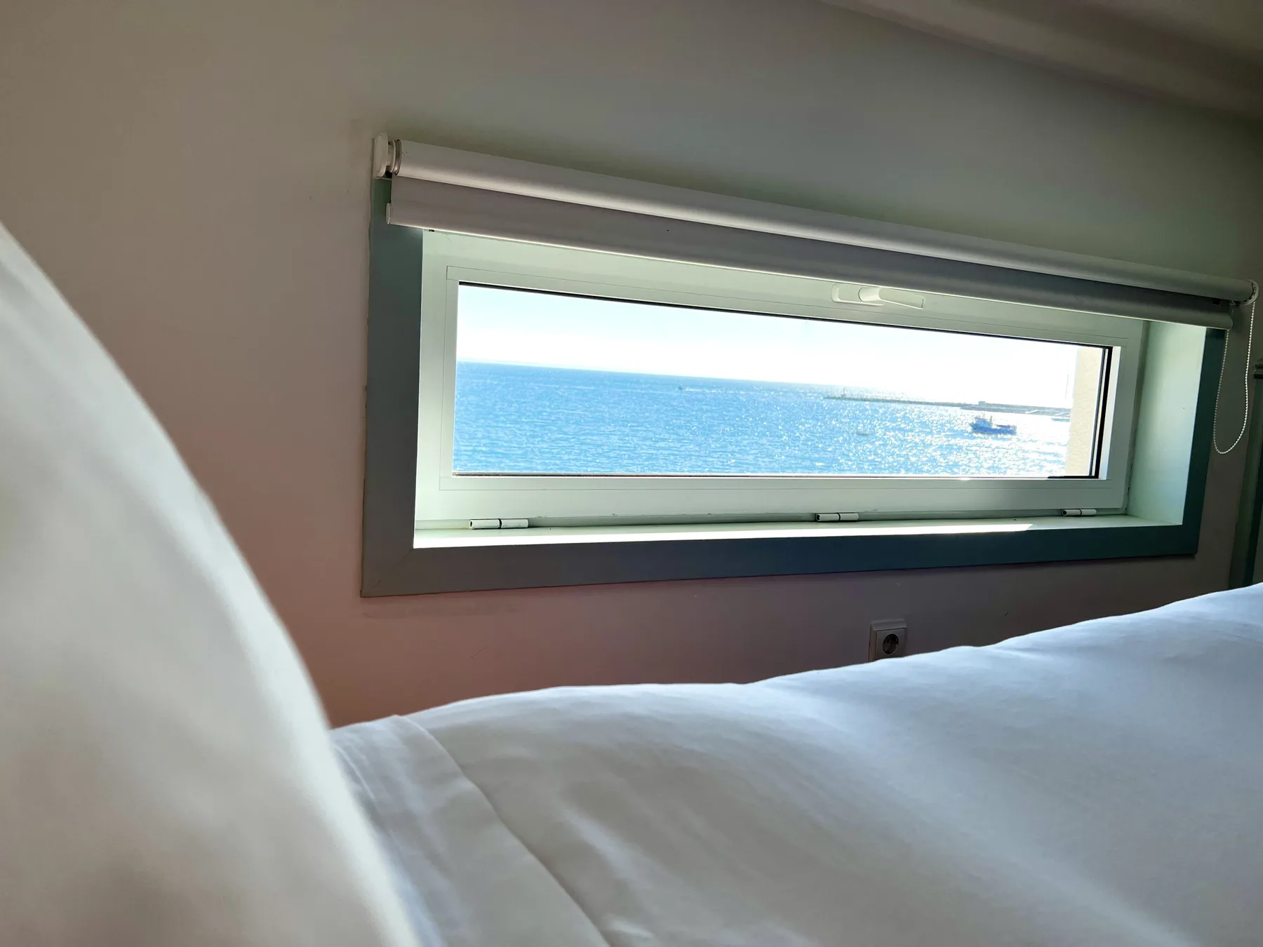 A bedroom with white bedding and a narrow window overlooking the sea.