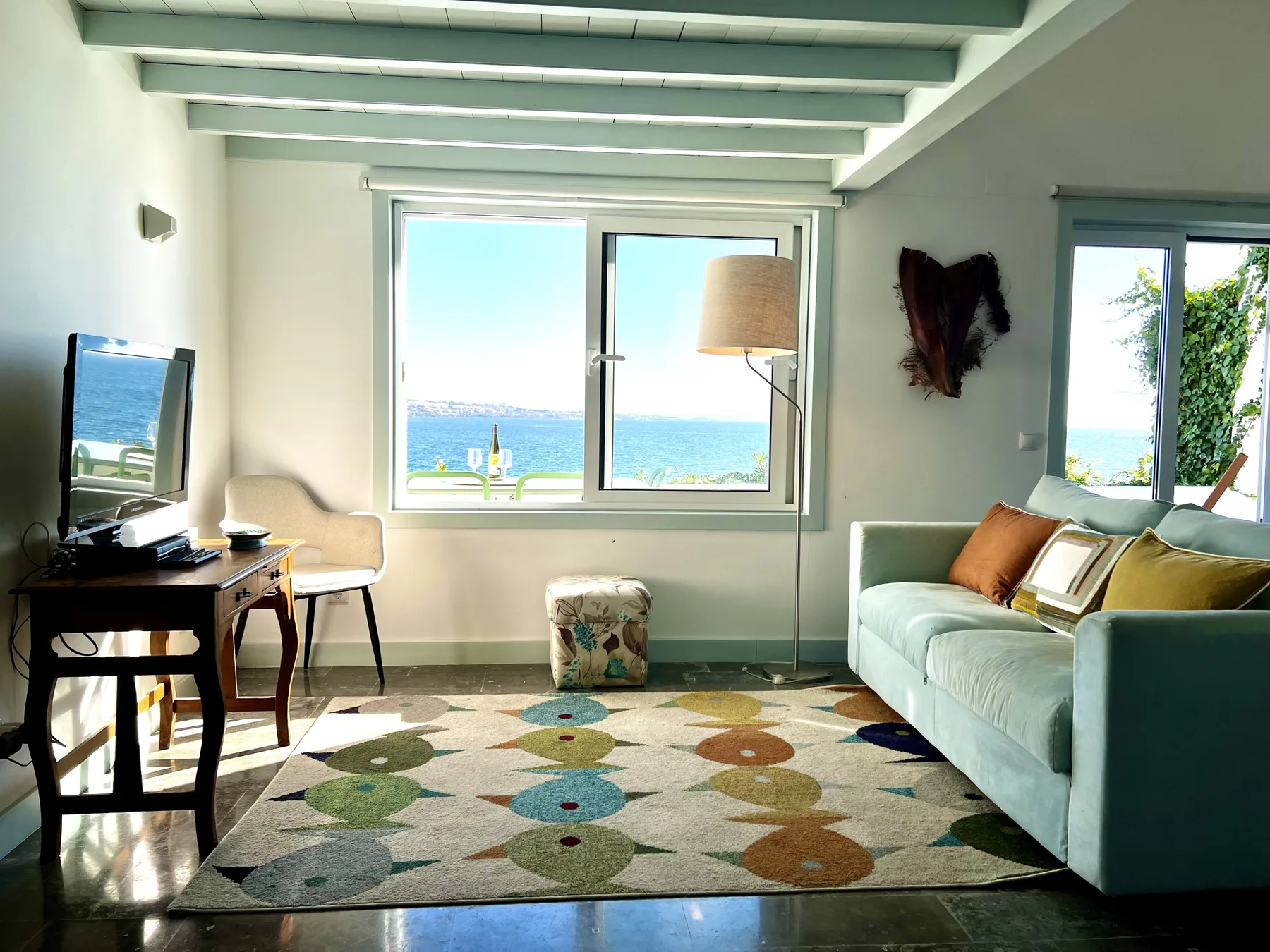 A bright living room with seating, a rug, and large windows overlooking the sea.