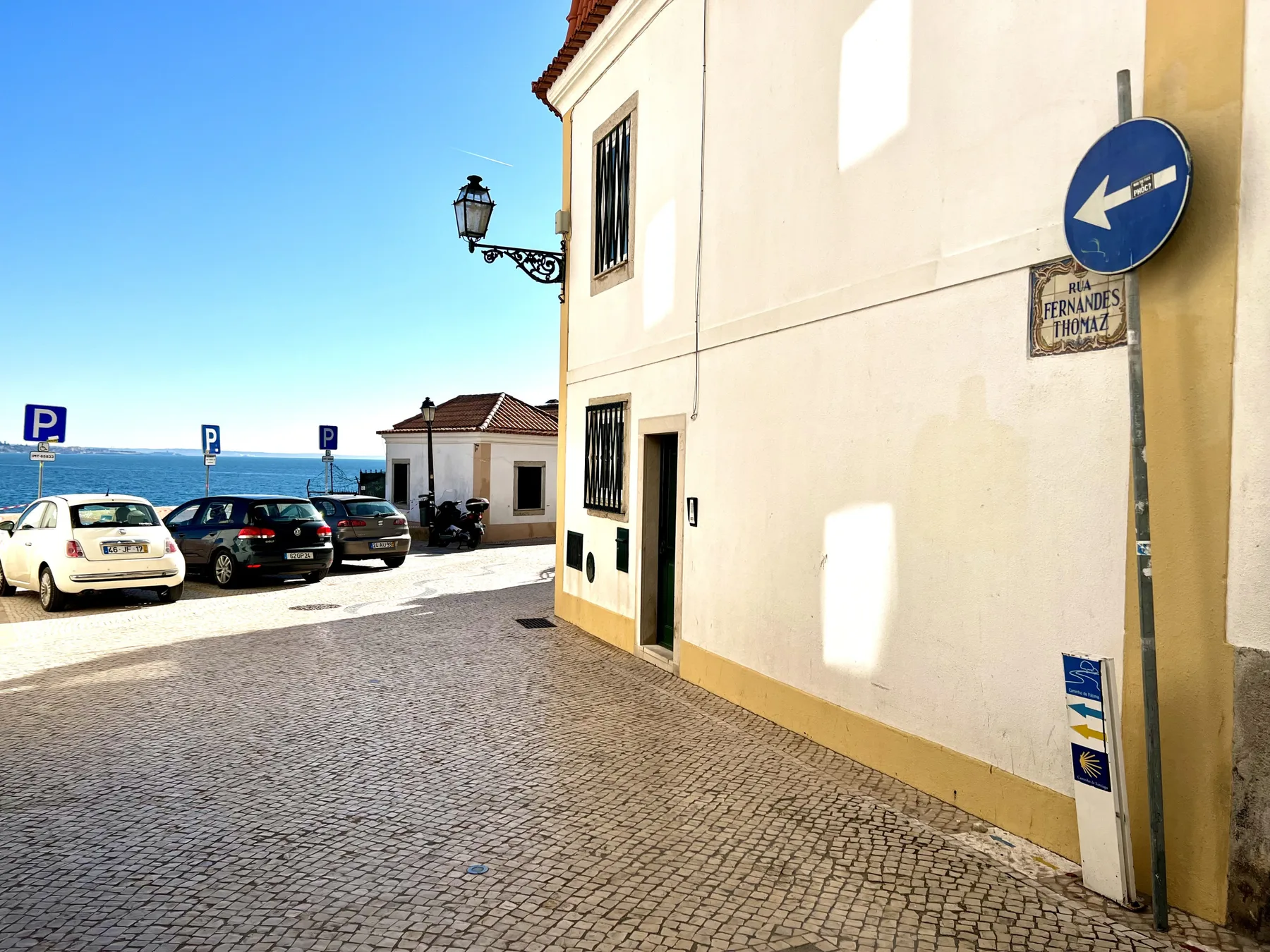 A cobbled street runs past a pale building, with parked cars and the sea beyond.
