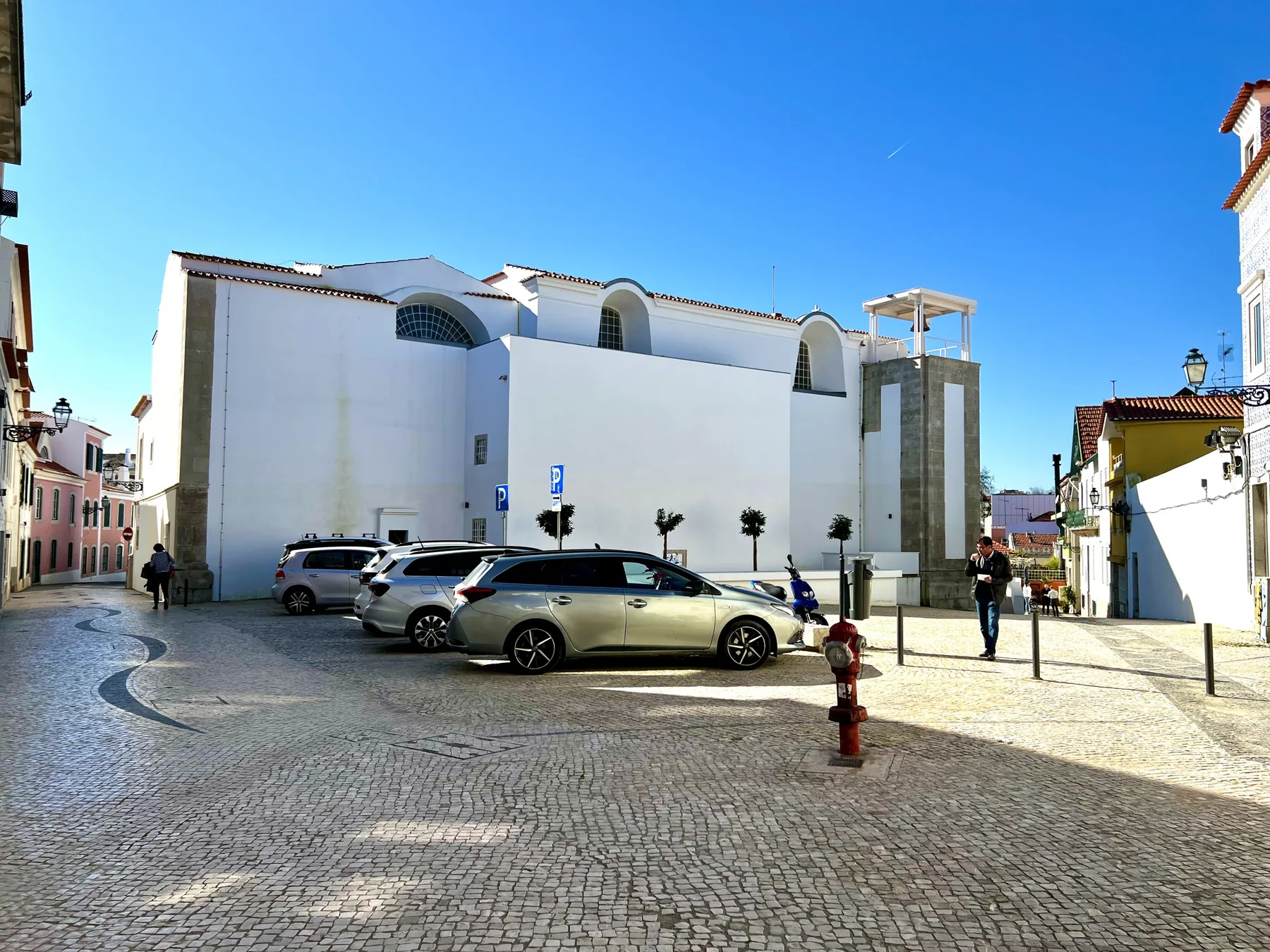 A cobblestone plaza with parked cars in front of a large white building.