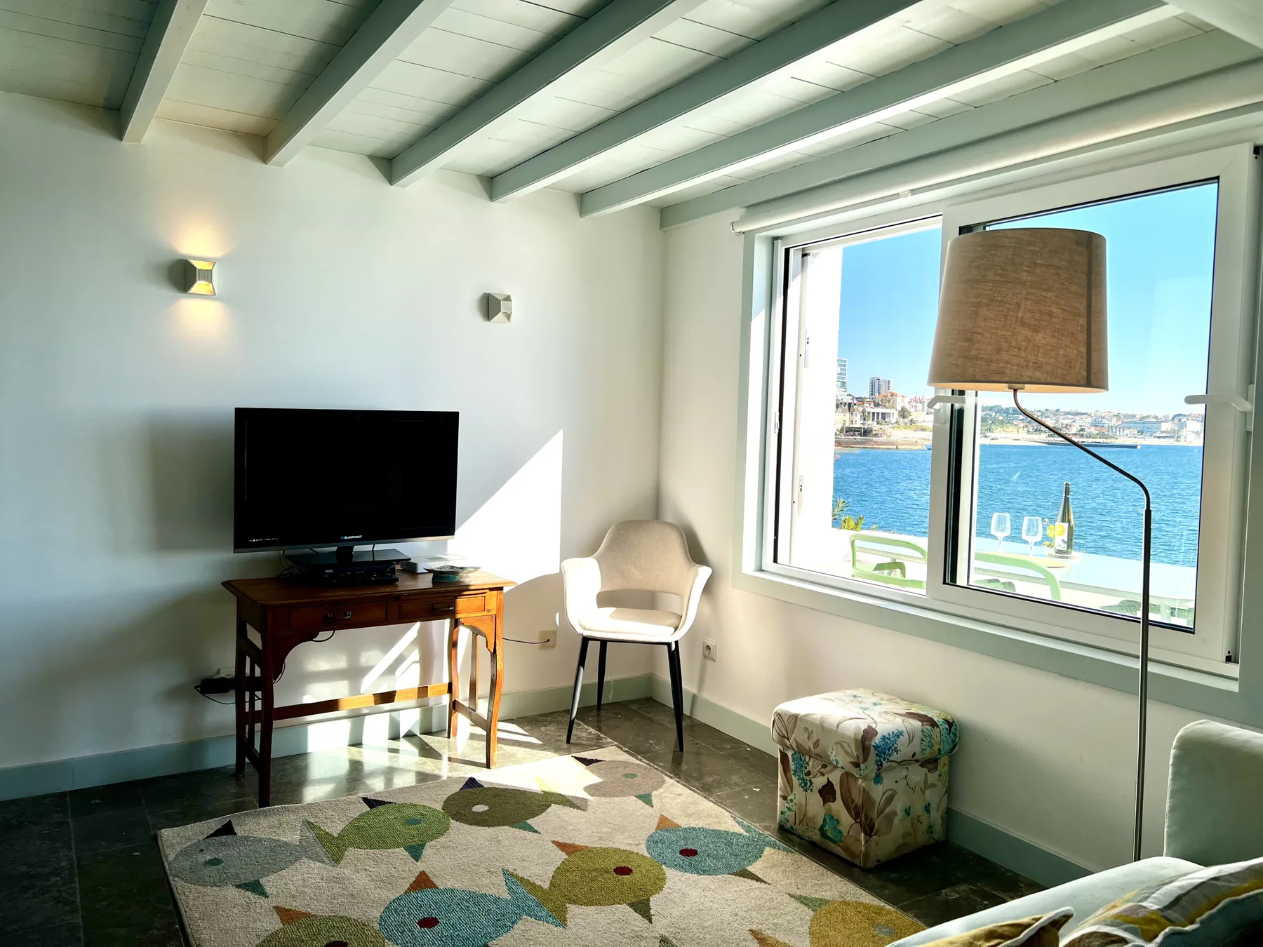 A bright living area has a television, seating, and a large window overlooking the water.