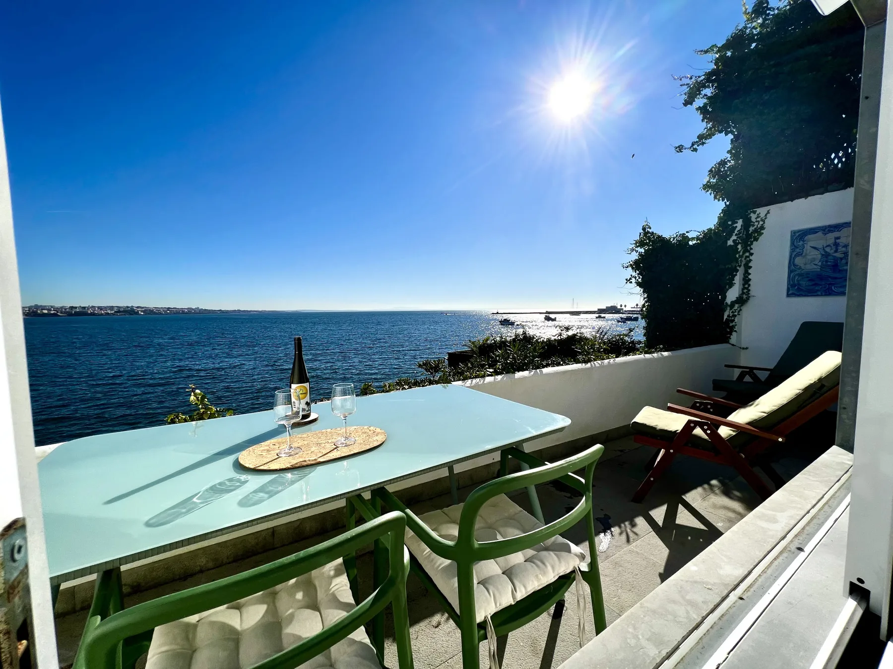 A seaside terrace overlooks the water, with a table and seating in the sun.