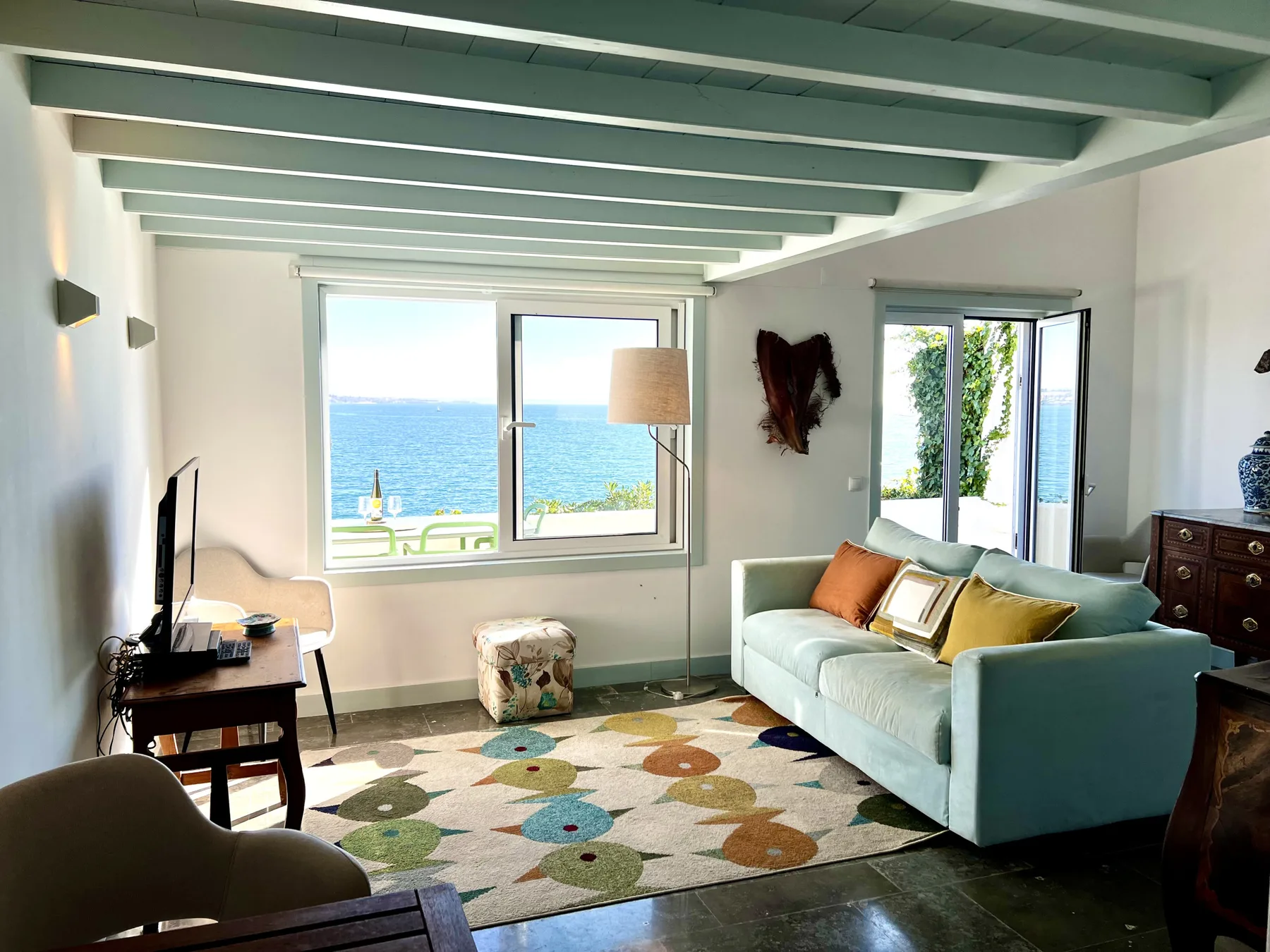 A bright living room with seating, a television, and a view of the sea.