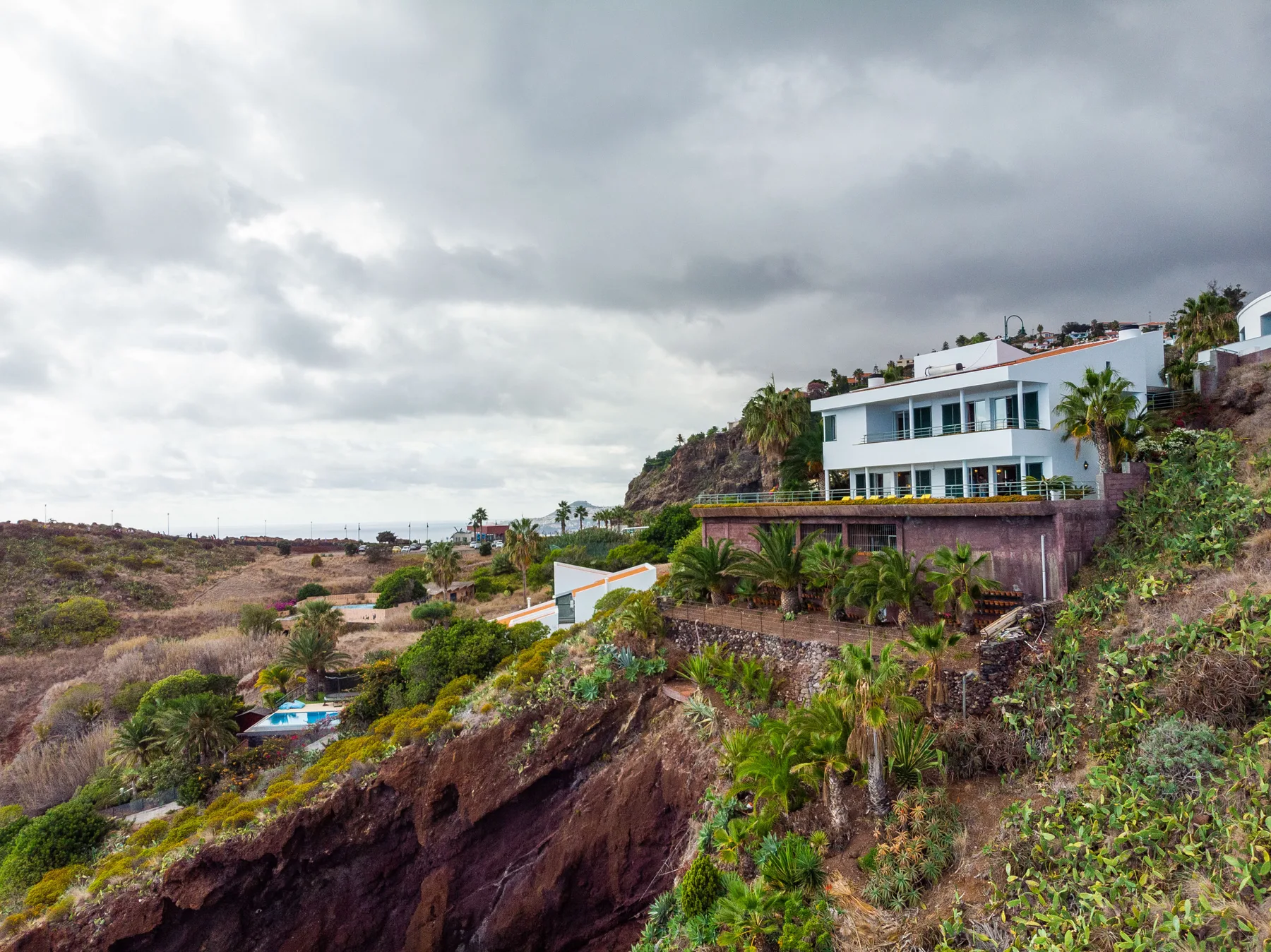 Una casa blanca de varias plantas se encuentra en una ladera con vegetación y cielo nublado.
