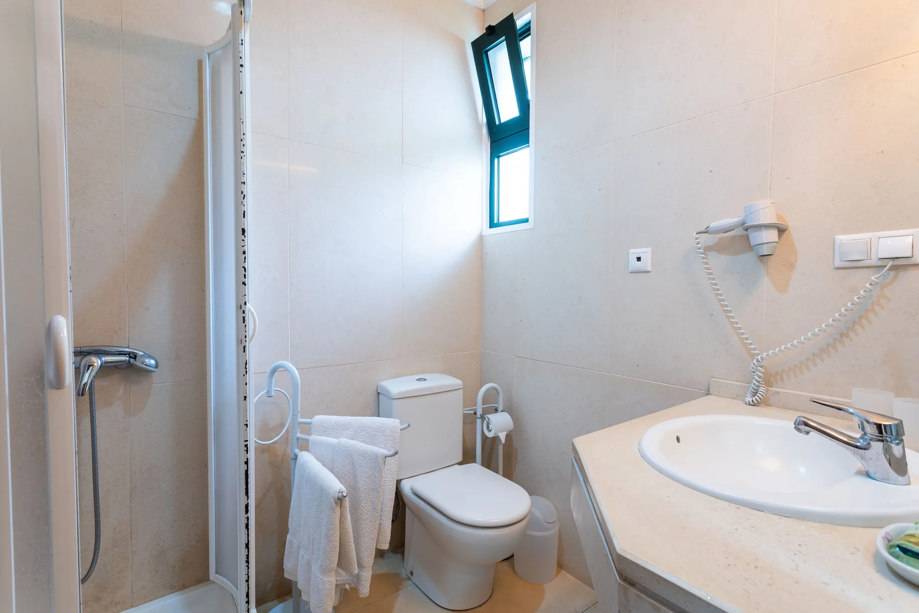 Un baño con azulejos, ducha, inodoro y lavabo bajo una ventana pequeña.