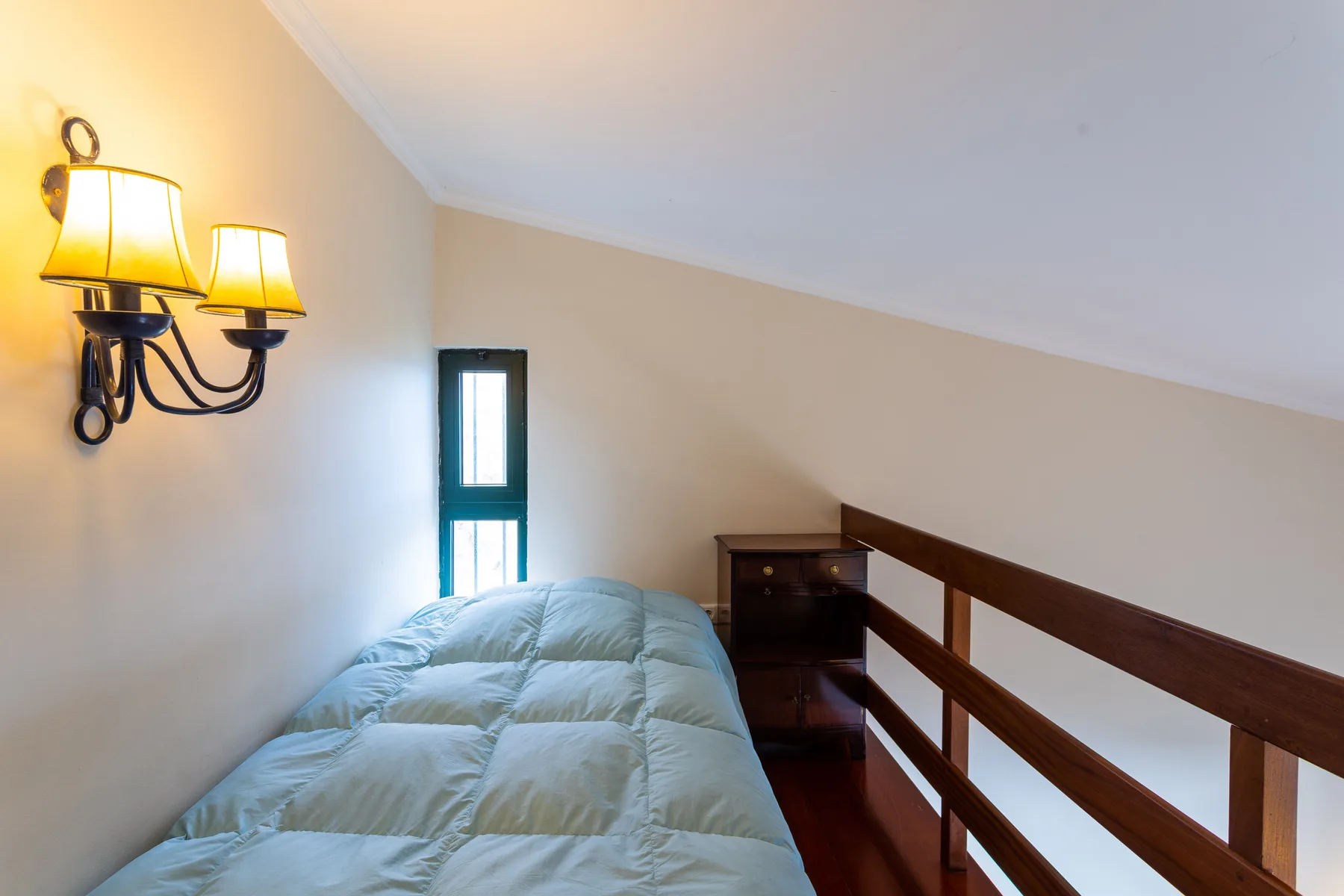 Zona de descanso en un altillo con una cama junto a una barandilla de madera y una ventana estrecha.