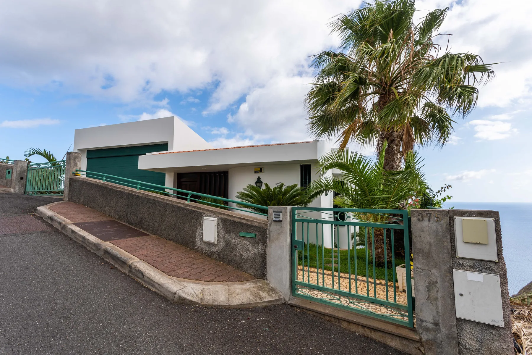 Una casa moderna tras un muro bajo y una puerta verde, con una palmera al lado.