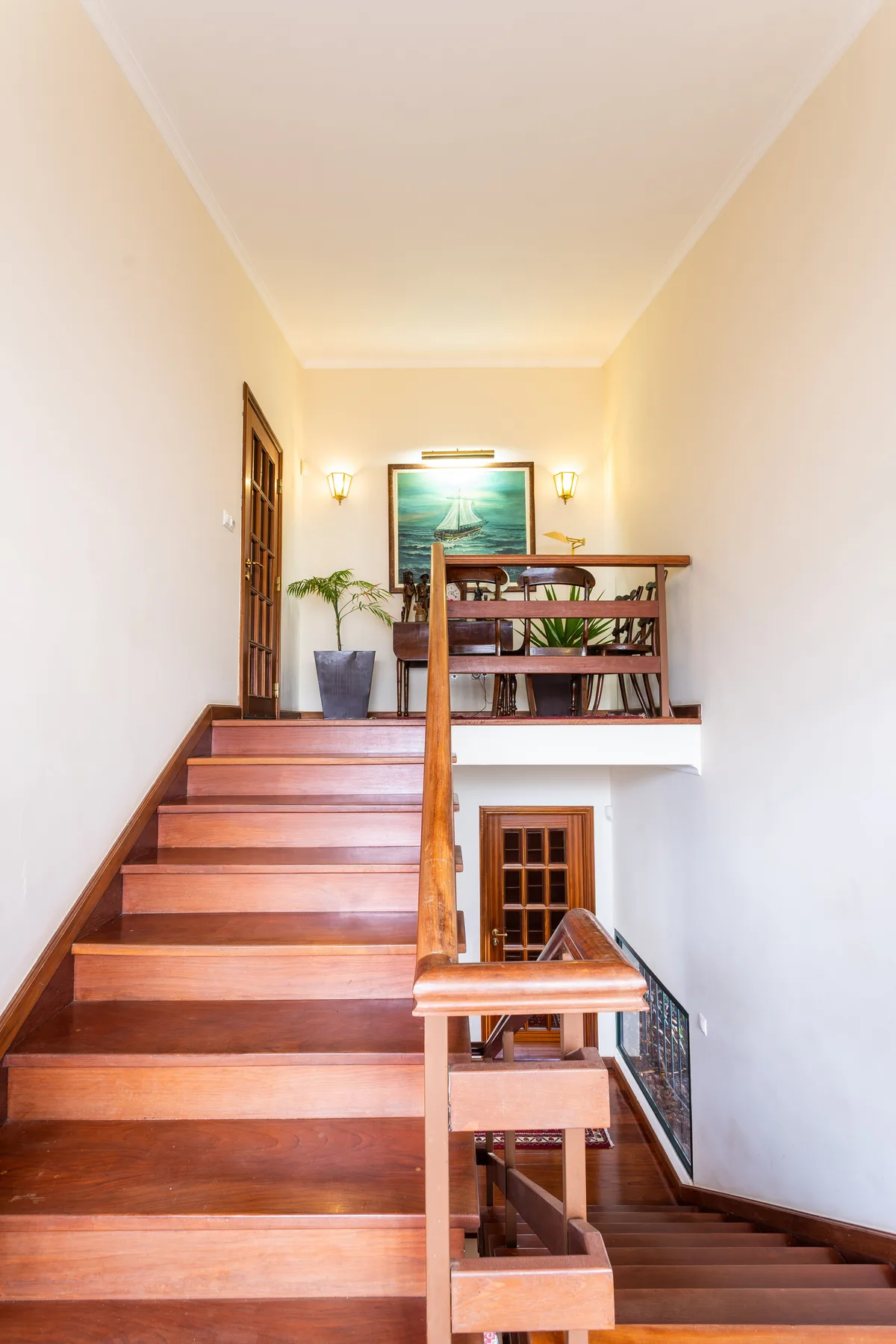 Una escalera de madera sube a un rellano con plantas y una obra enmarcada.