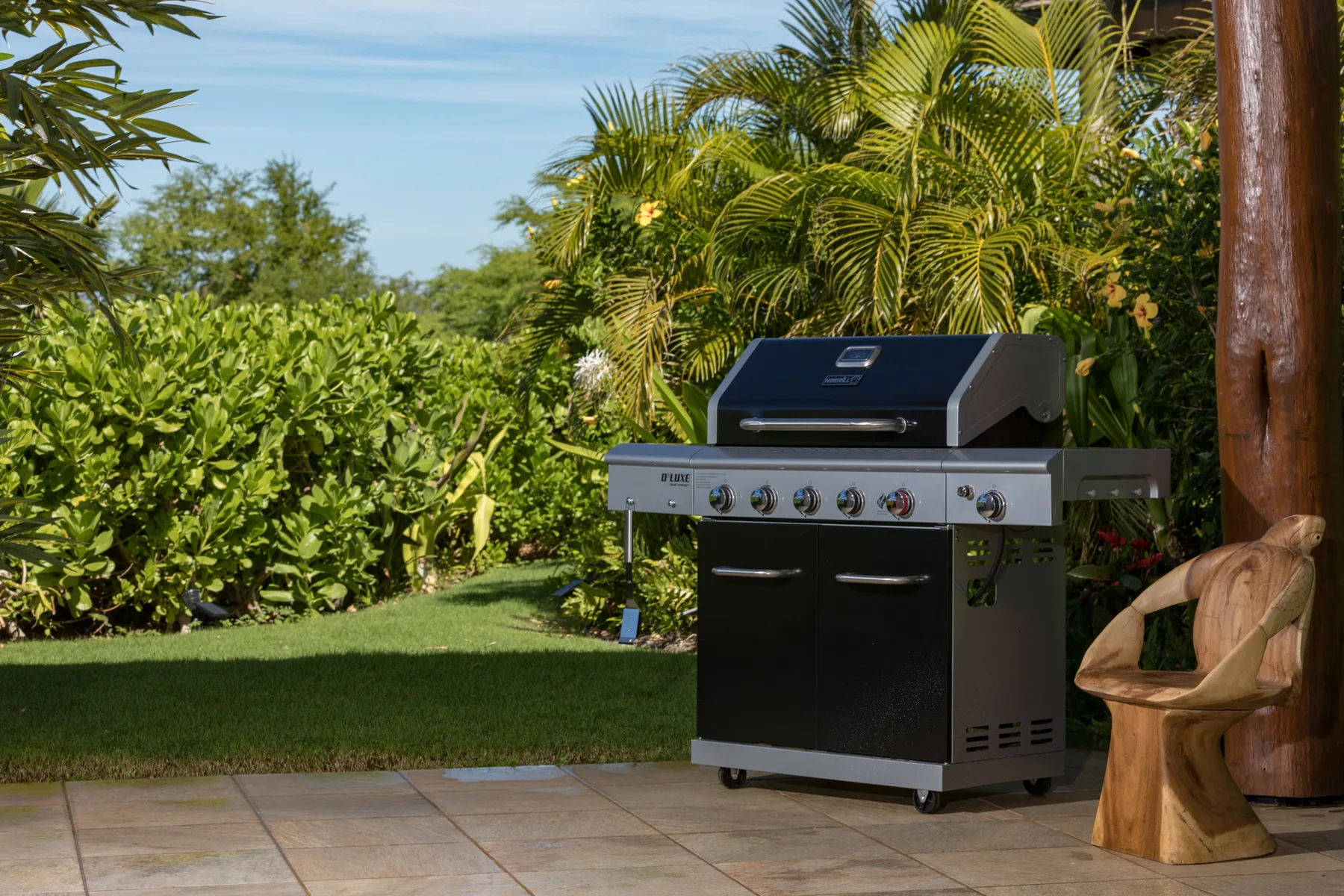 Una parrilla al aire libre está en un patio embaldosado junto a una silla de madera y abundante vegetación.