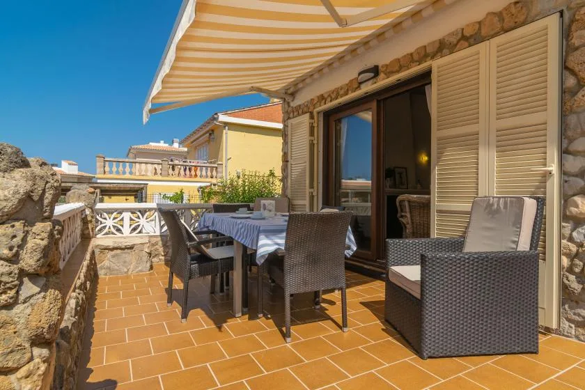 A sunny terrace has a dining table and seating beneath a striped awning.