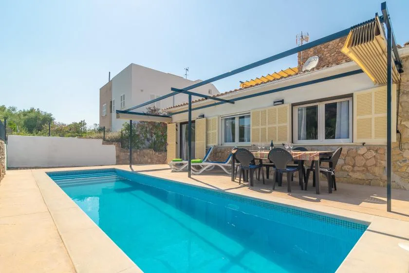 A swimming pool beside a house with outdoor seating under a pergola.