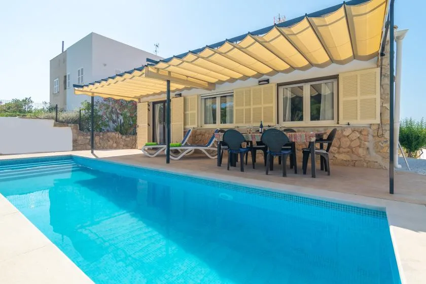 An outdoor swimming pool sits beside a patio with seating under a shade canopy.