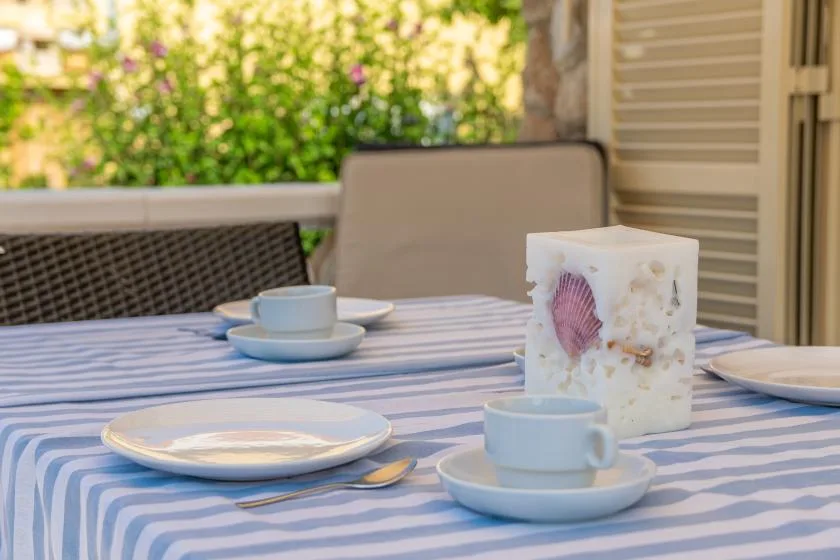 An outdoor dining table is set with cups and plates beside a leafy garden.