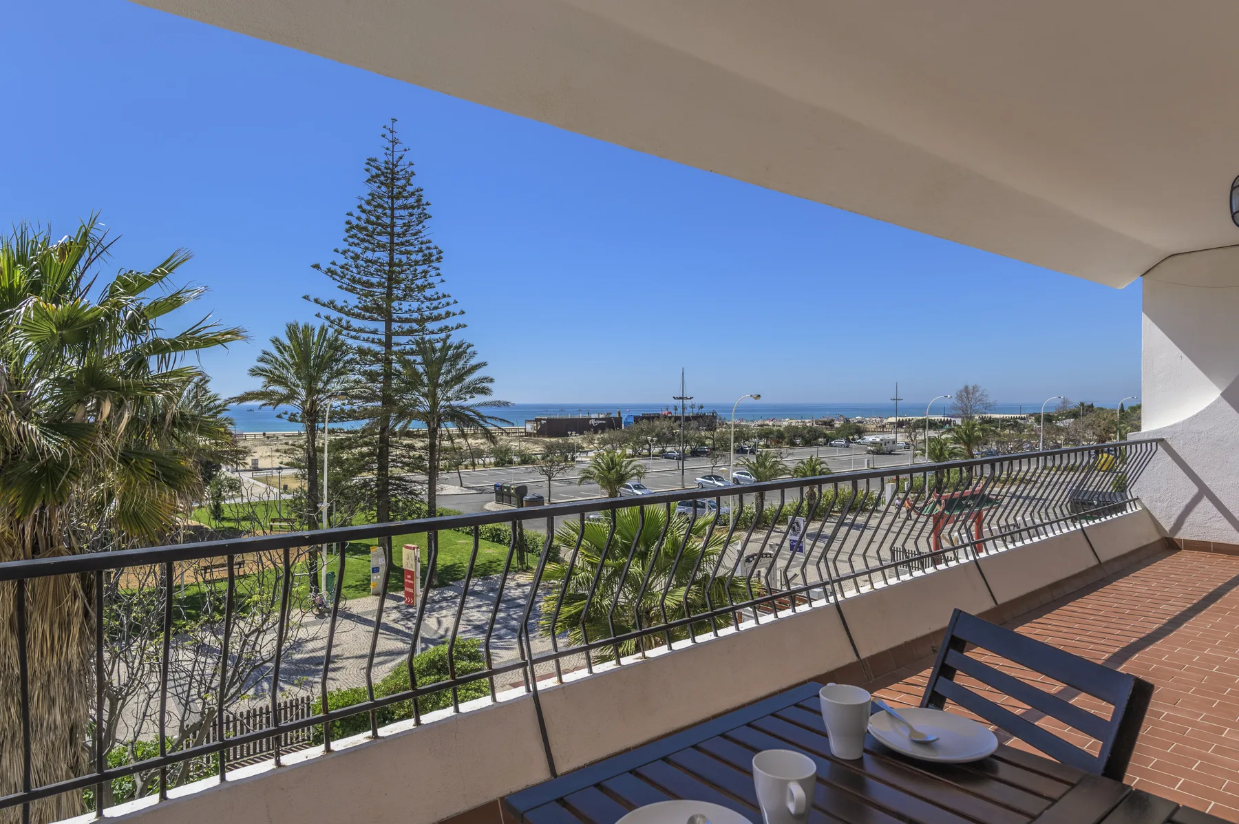 A balcony overlooks palm trees, a coastal path, and the sea.