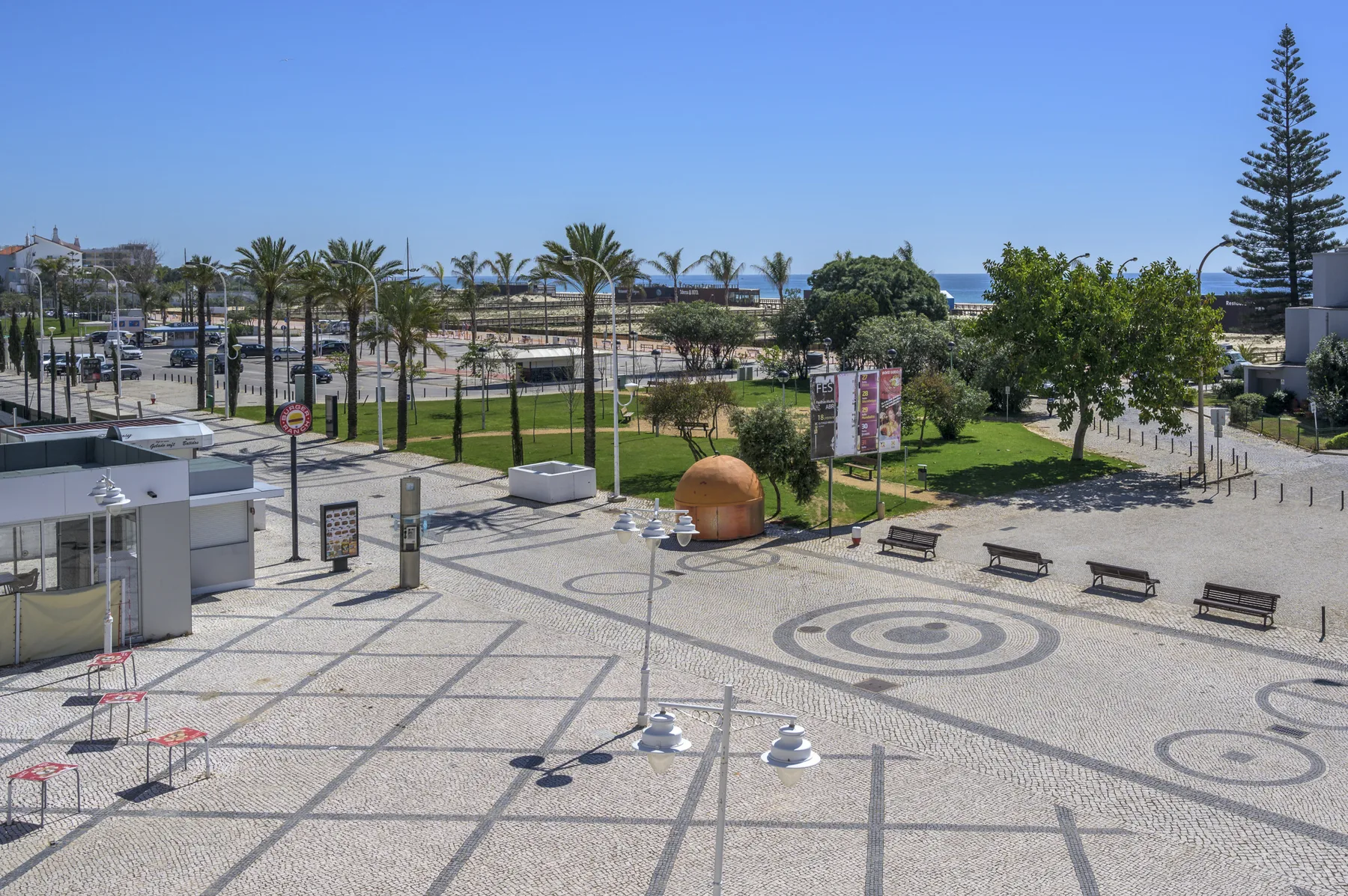 A paved plaza with benches, palm trees, and a strip of ocean in the background.