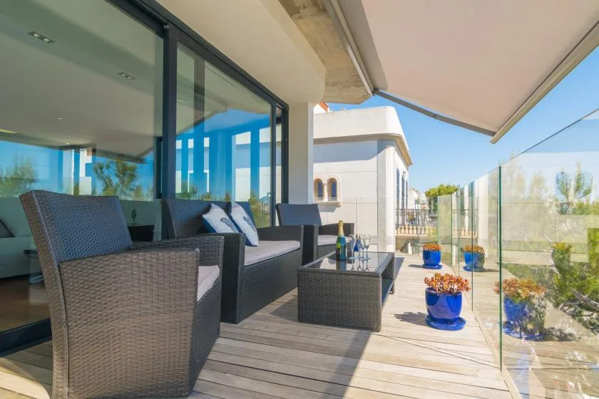 A furnished balcony with outdoor seating and potted plants beside a glass railing.