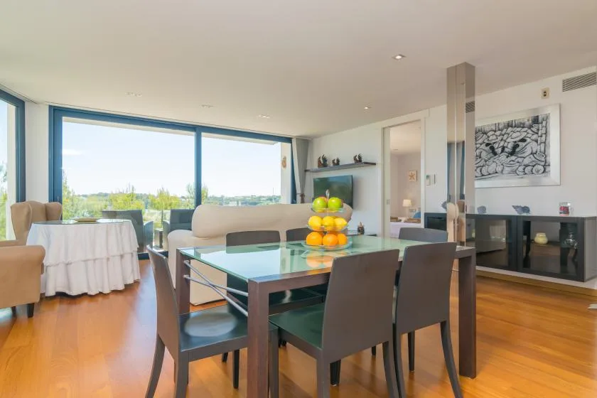 A dining area with a glass-top table and large windows overlooking greenery.