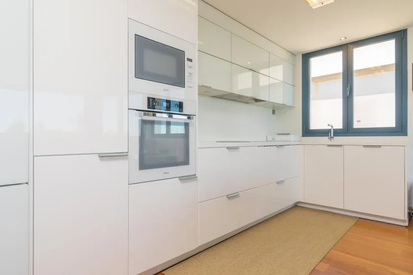 A bright modern kitchen with white cabinetry, built-in appliances, and a large window.