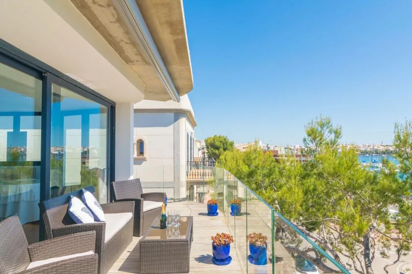 A sunny balcony with seating and potted plants overlooks trees and the sea.