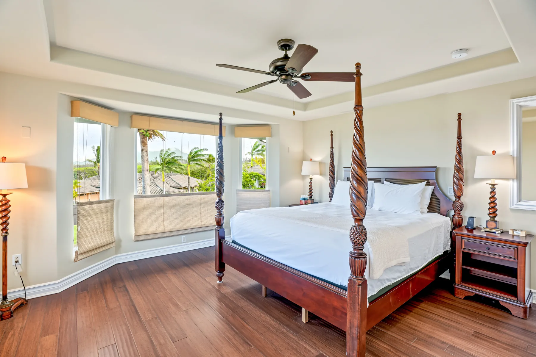 A bedroom with a large wooden bed, white bedding, and windows overlooking palm trees.
