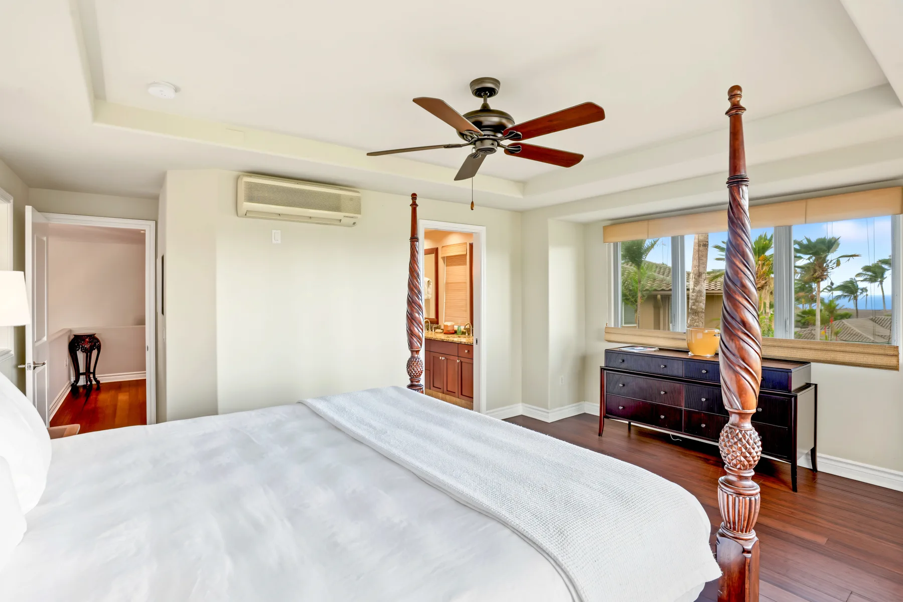 A bright bedroom with a large bed, ceiling fan, and windows overlooking palm trees.