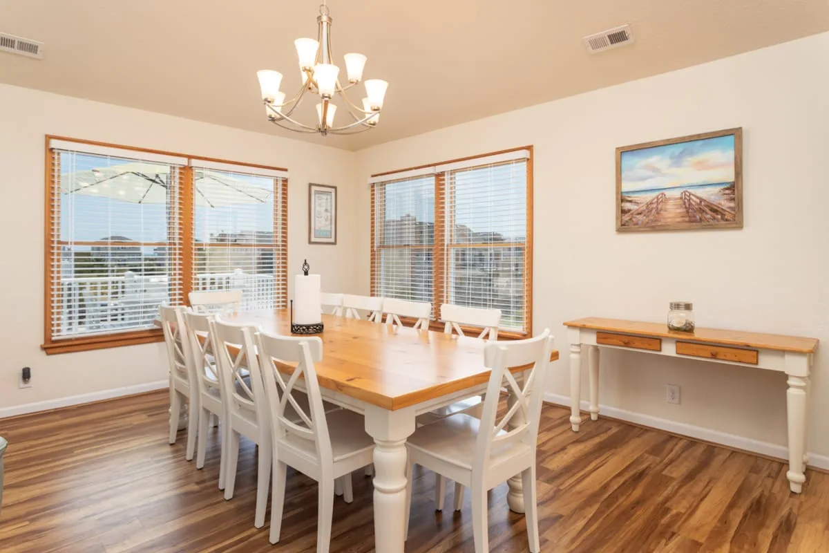 A dining room with a wooden table, white chairs, and large windows.