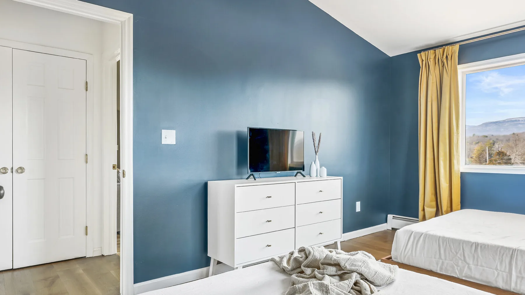 Un dormitorio con una cama, una cómoda blanca y un televisor frente a una pared azul.