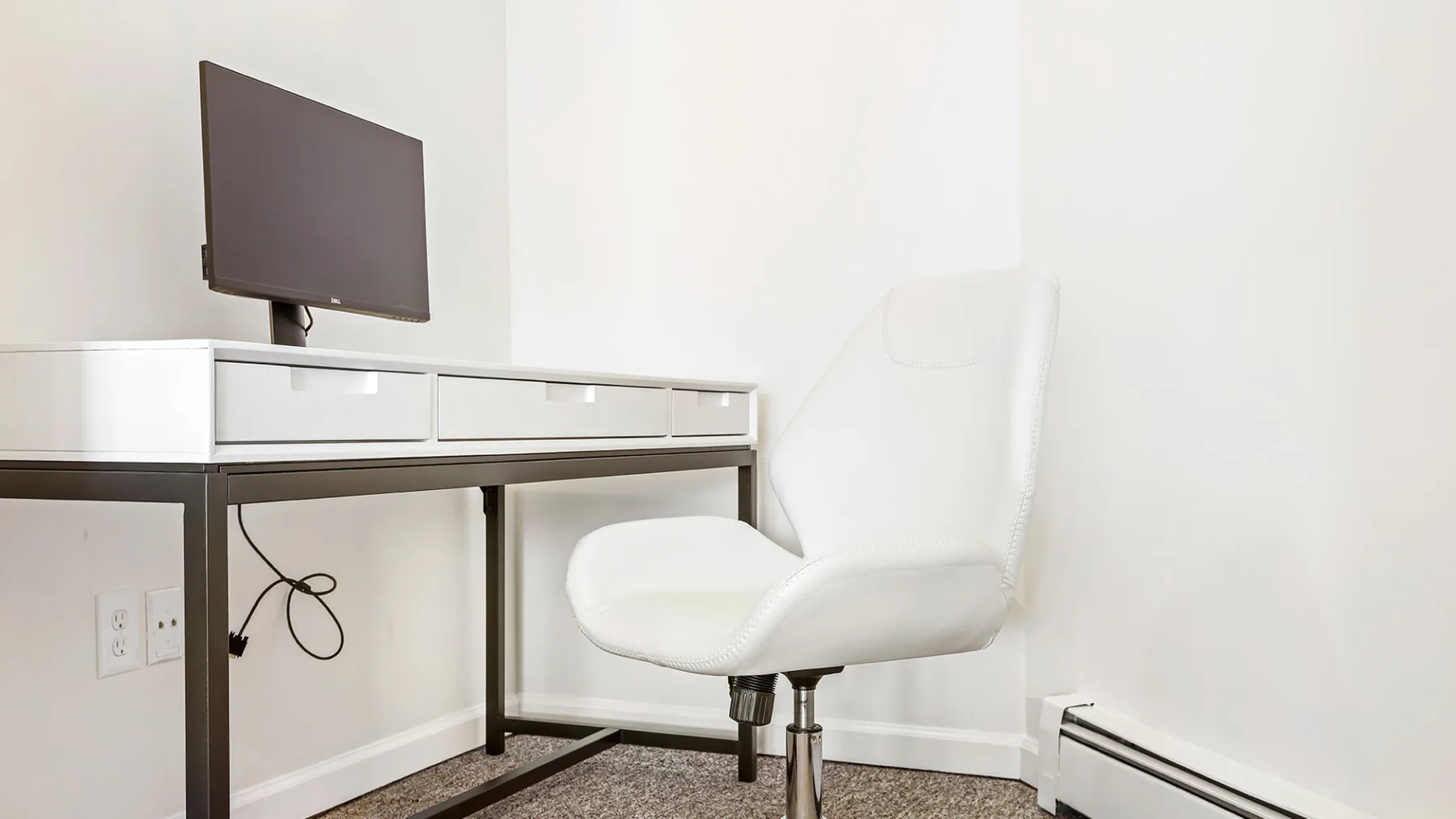 Un escritorio con monitor y una silla blanca en una habitación luminosa.