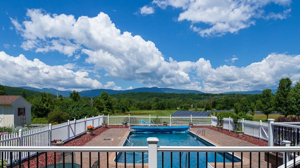Una piscina al aire libre está rodeada por una terraza cercada, con colinas verdes a lo lejos.